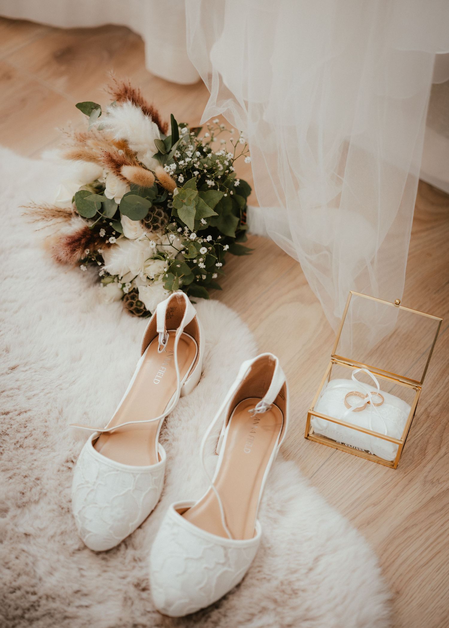 White lace bridal heels beside a wildflower bouquet, wedding rings in a glass box, on a fluffy white rug.