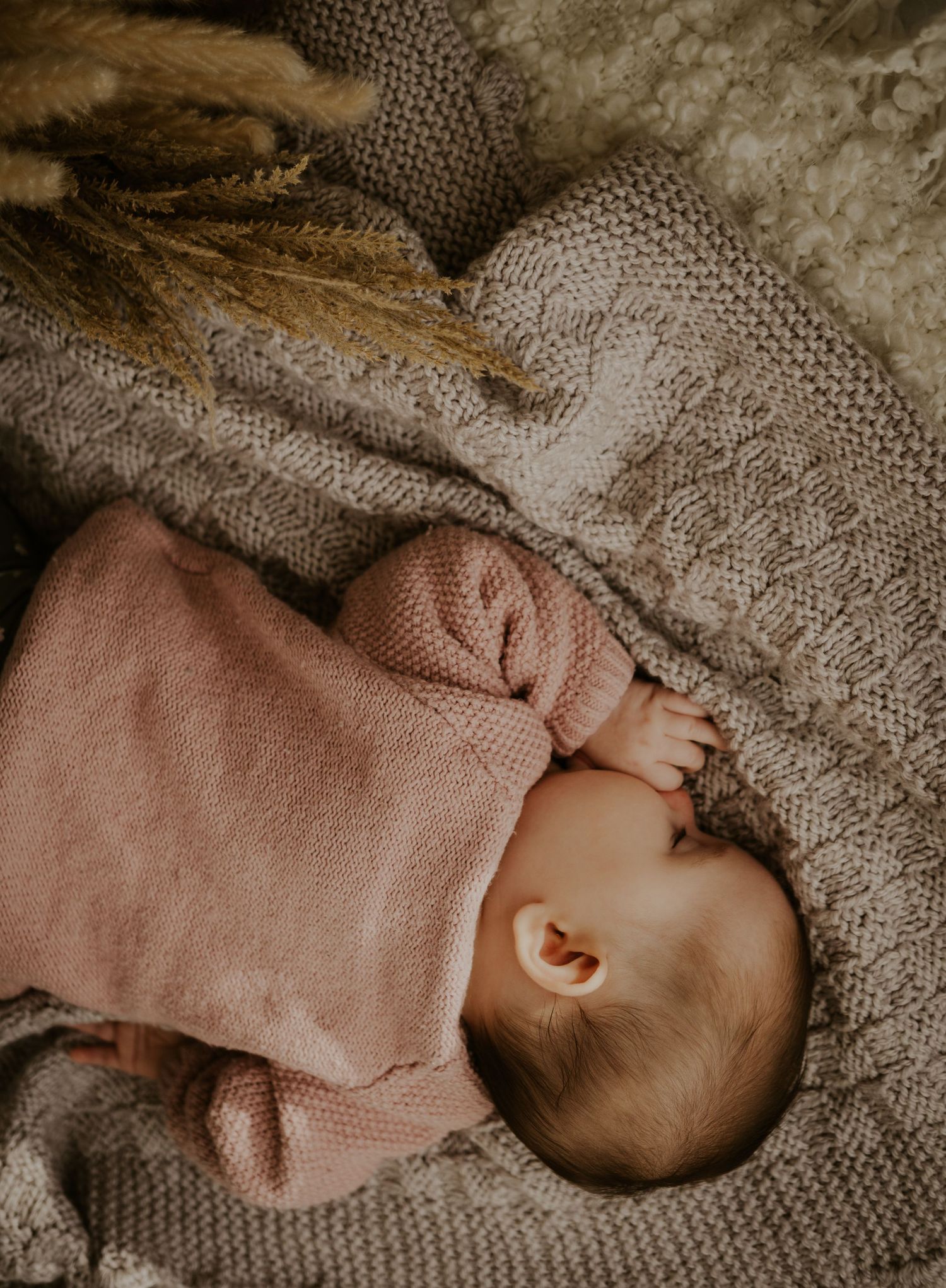 A sleeping baby in a cozy knit sweater rests peacefully on a textured gray blanket in natural lighting.