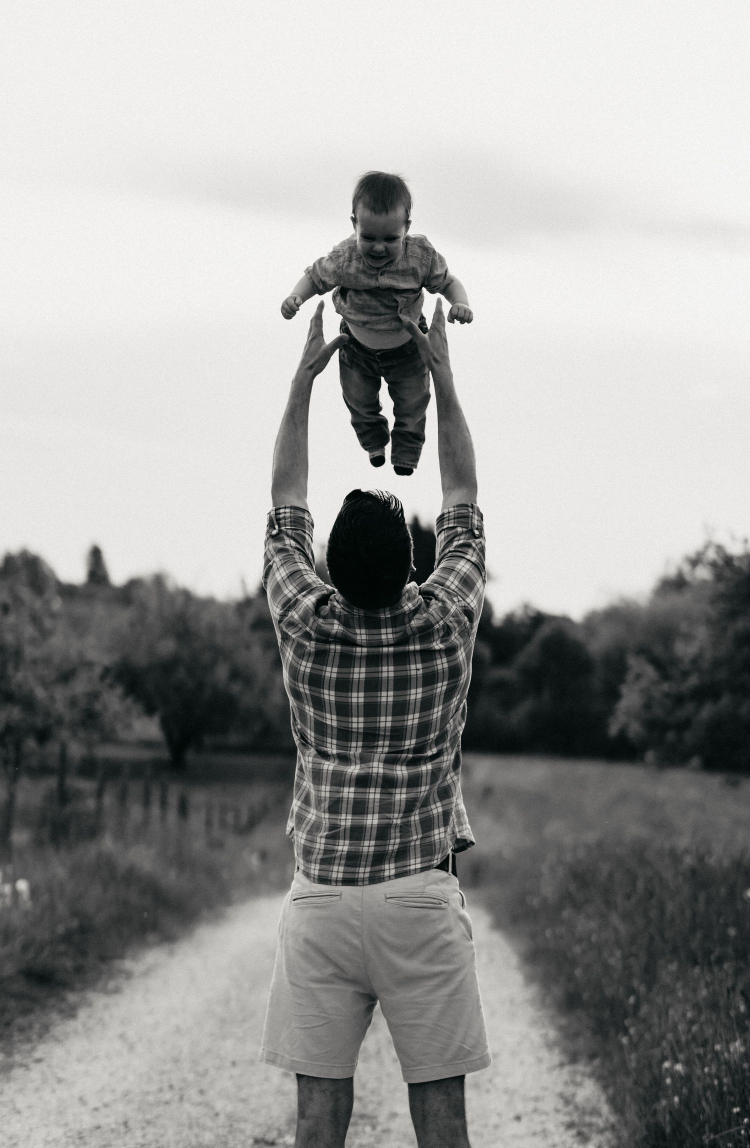 A sequence of black and white photos showing a person in a plaid shirt tossing a child up in the air on a rural path.