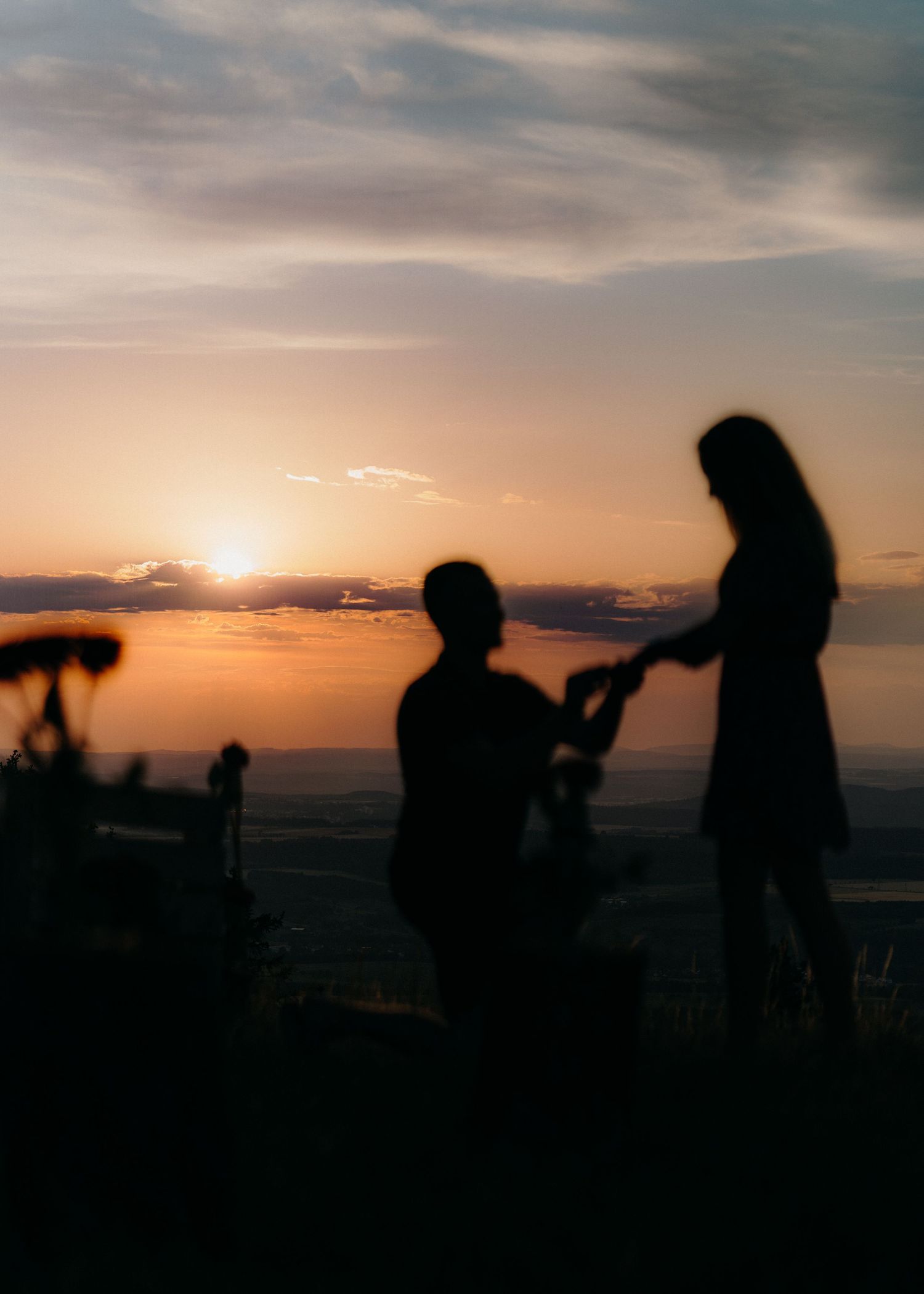 Two silhouettes of people against a dramatic sunset sky with one figure standing and one kneeling during a special moment.