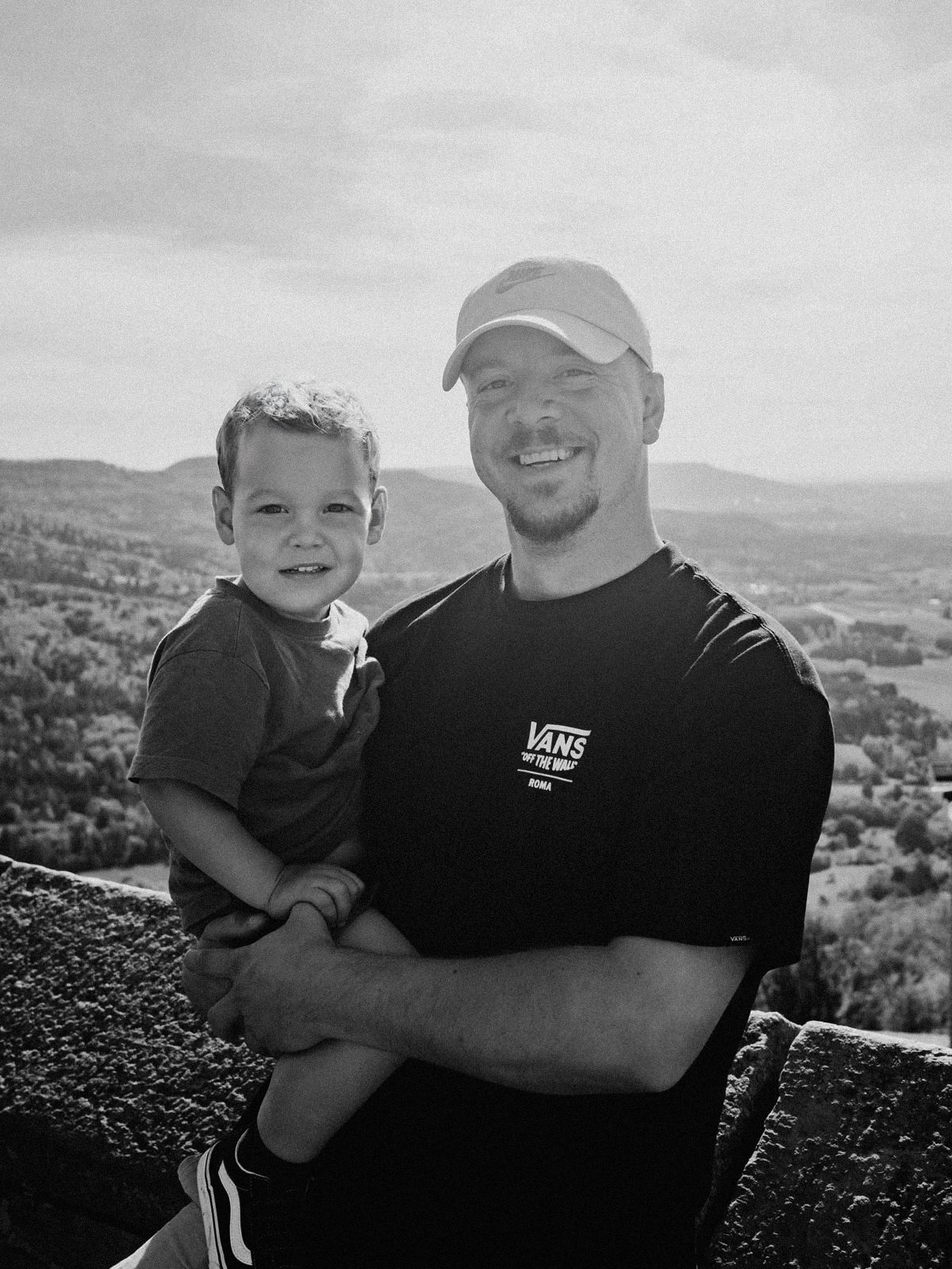 Man in Vans t-shirt and cap holds smiling toddler on scenic overlook with rolling hills backdrop.