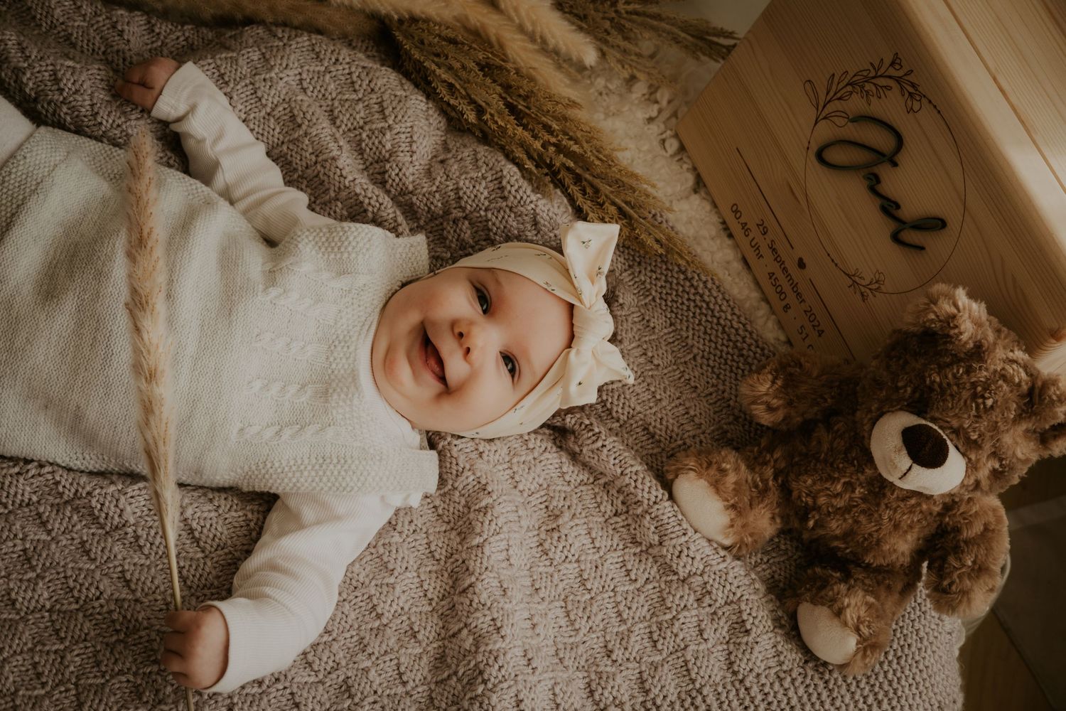 A smiling baby in white clothing lies on a knitted blanket next to a brown teddy bear.