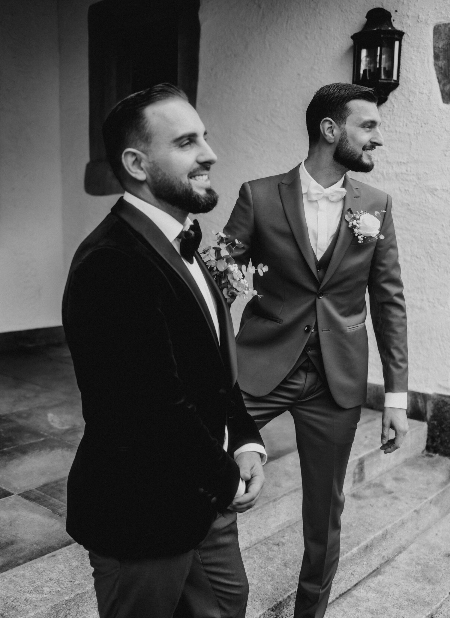 Two grooms in elegant suits share a joyful moment at their wedding, one in black velvet and one in gray.