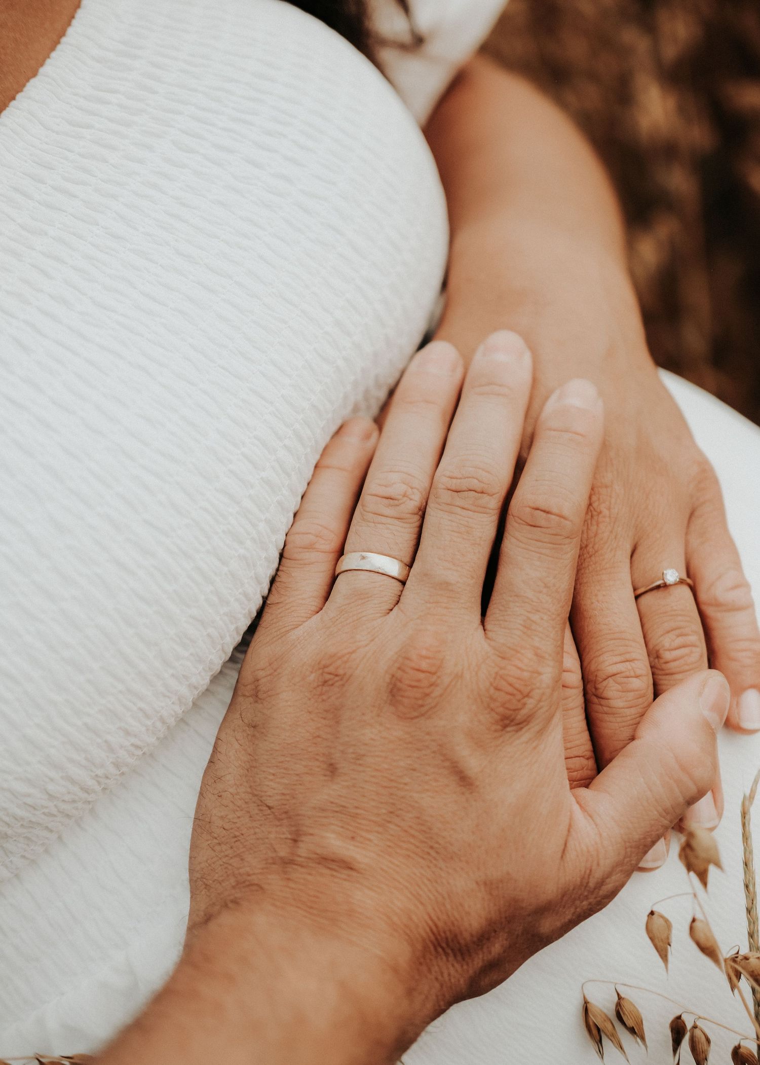 Two hands wearing wedding rings touch tenderly in a romantic moment against a white fabric background.