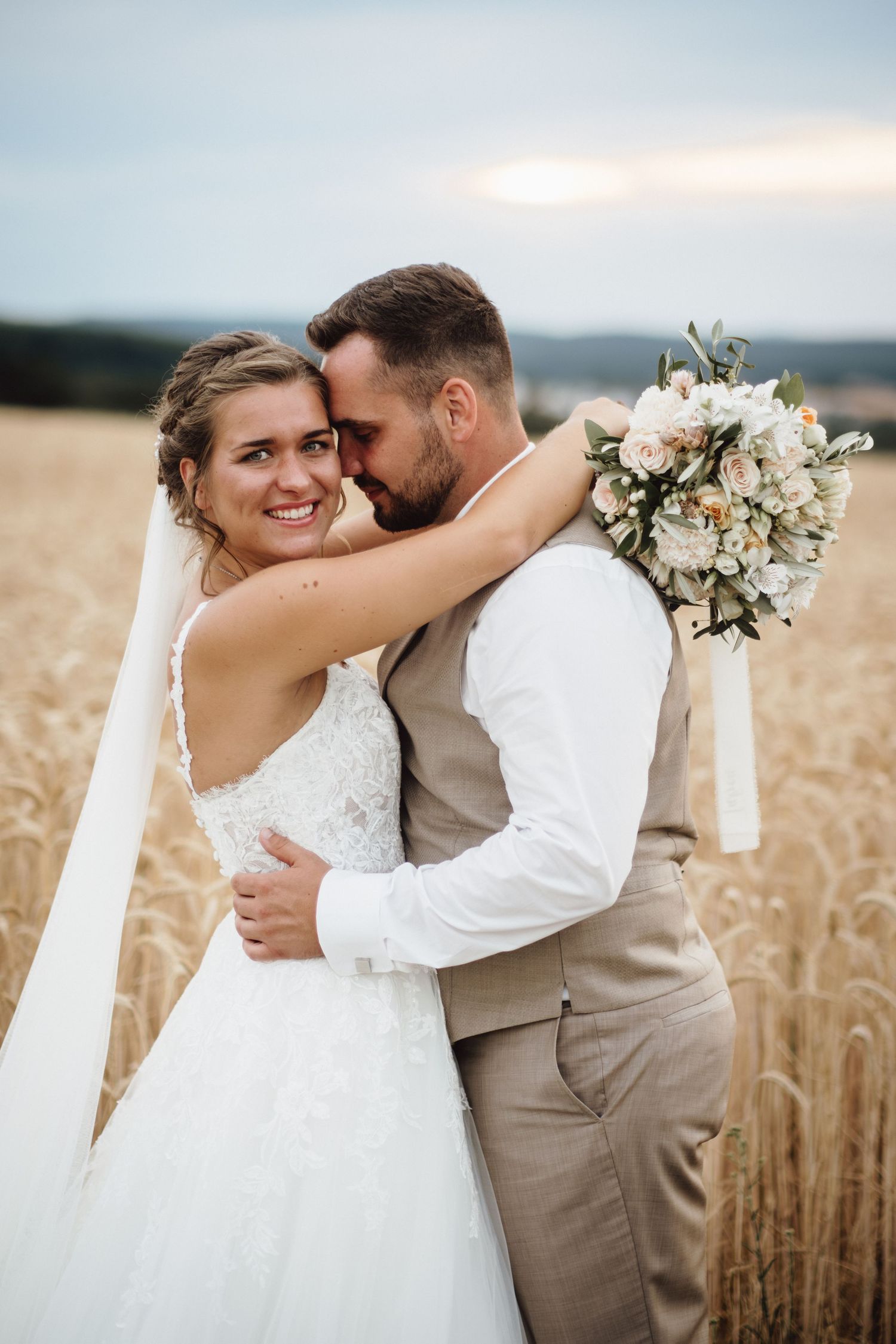Romantic wedding couple embraces in golden wheat field at sunset with white floral bouquet and flowing veil.