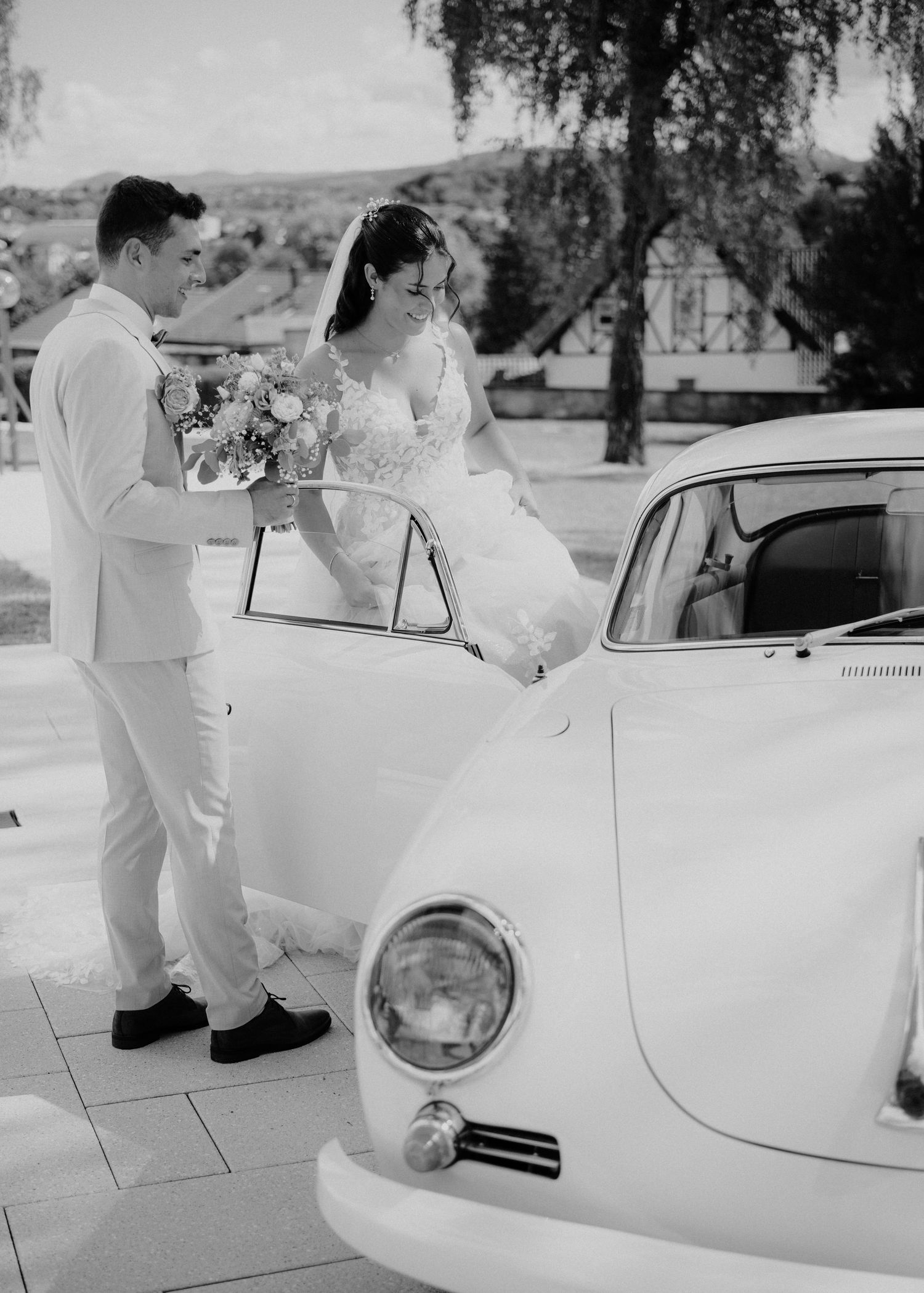 Bride and groom in white attire standing beside a vintage white car, holding a bouquet on their wedding day.