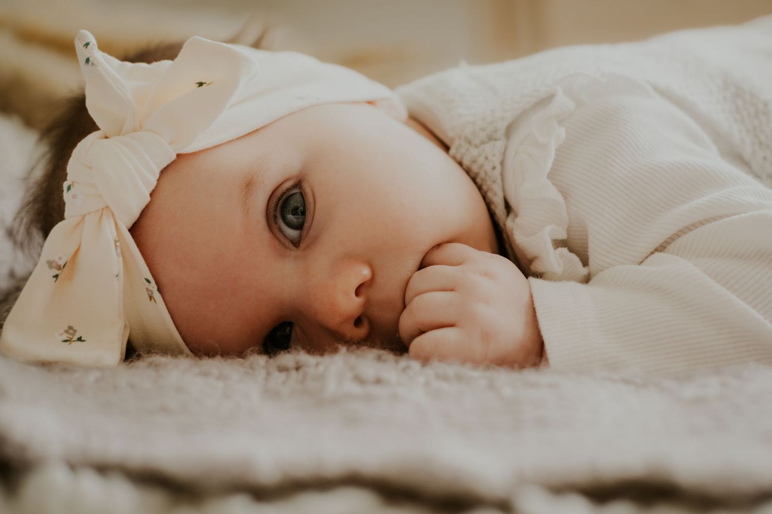 A baby wearing white clothing lies on white bedding while looking towards the camera.
