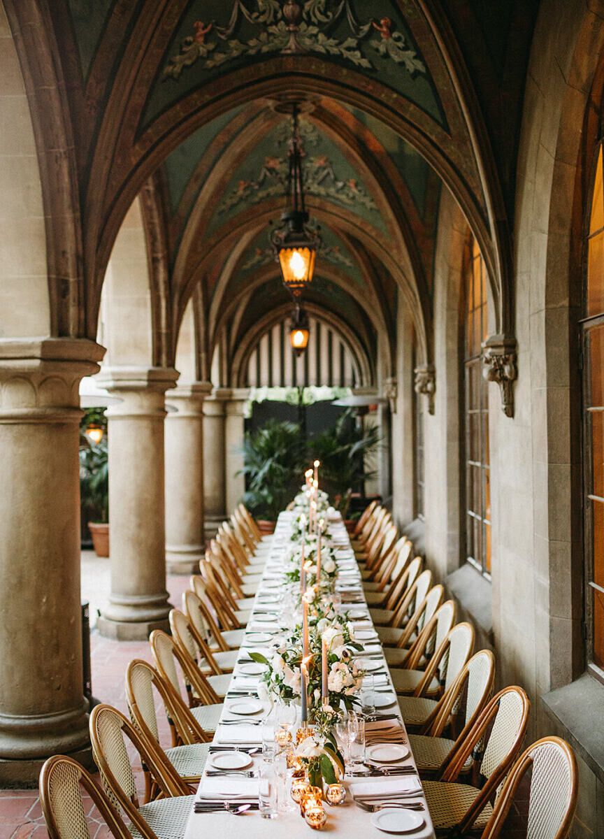 Long elegant dining table set under arched stone colonnade with warm lighting and white chairs for wedding reception.