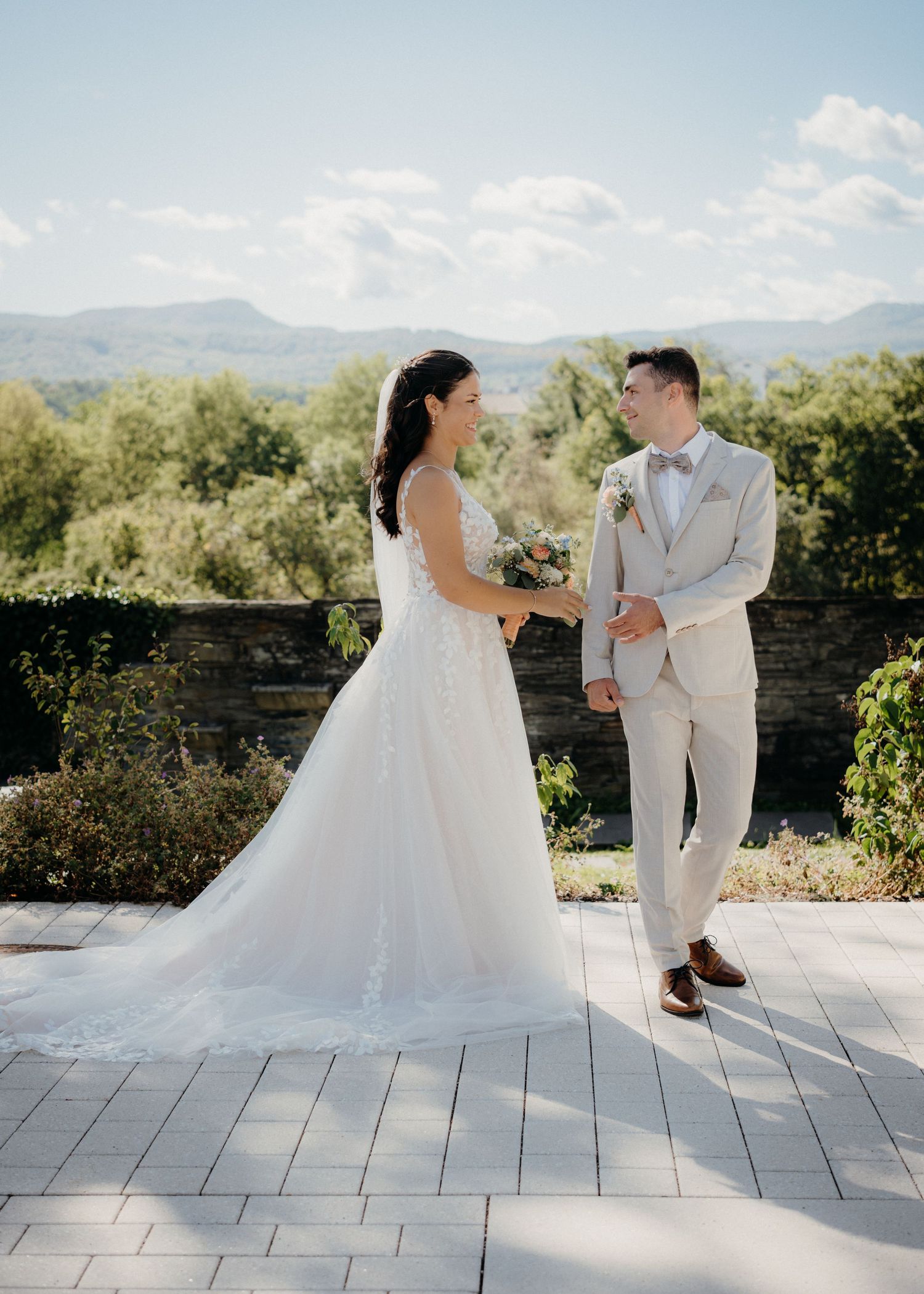Bride in white lace gown and groom in tan suit share a romantic first look on a sunny outdoor terrace.