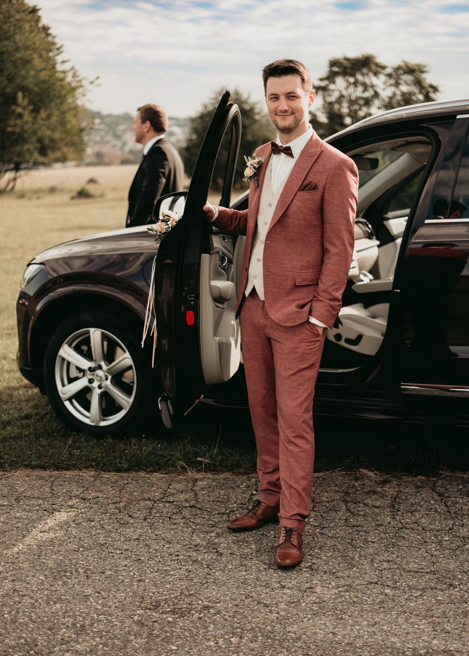 A person in a dusty rose colored suit stands next to a black luxury SUV on a dirt road in a rural setting.