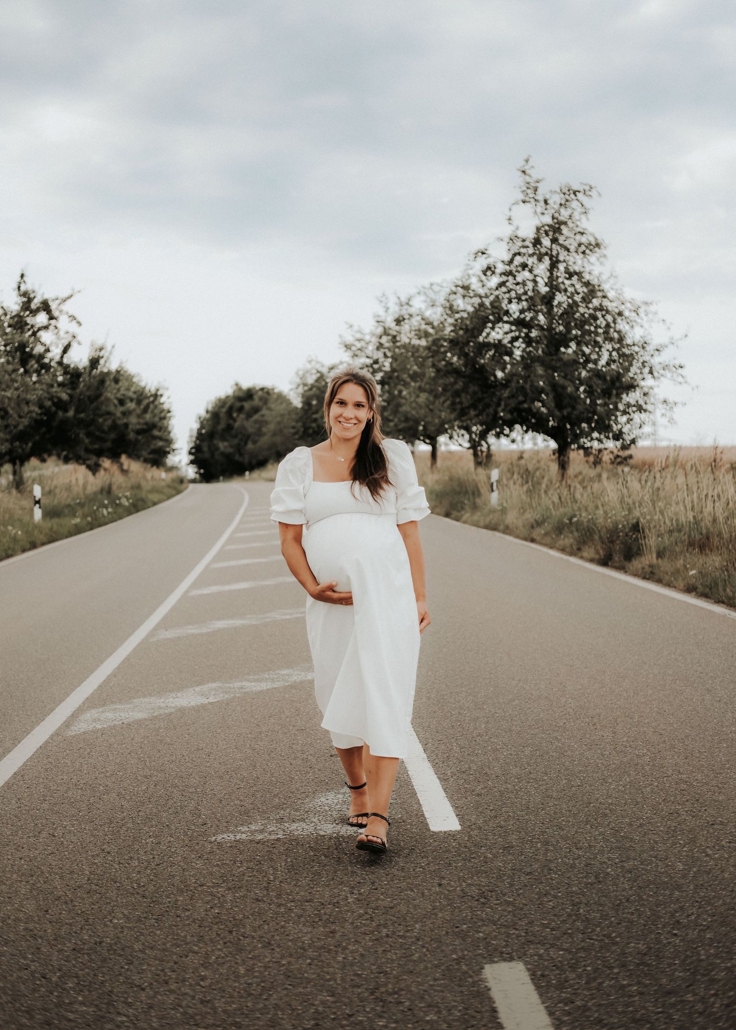 A person in a white dress twirls and dances playfully on an empty rural road surrounded by trees.