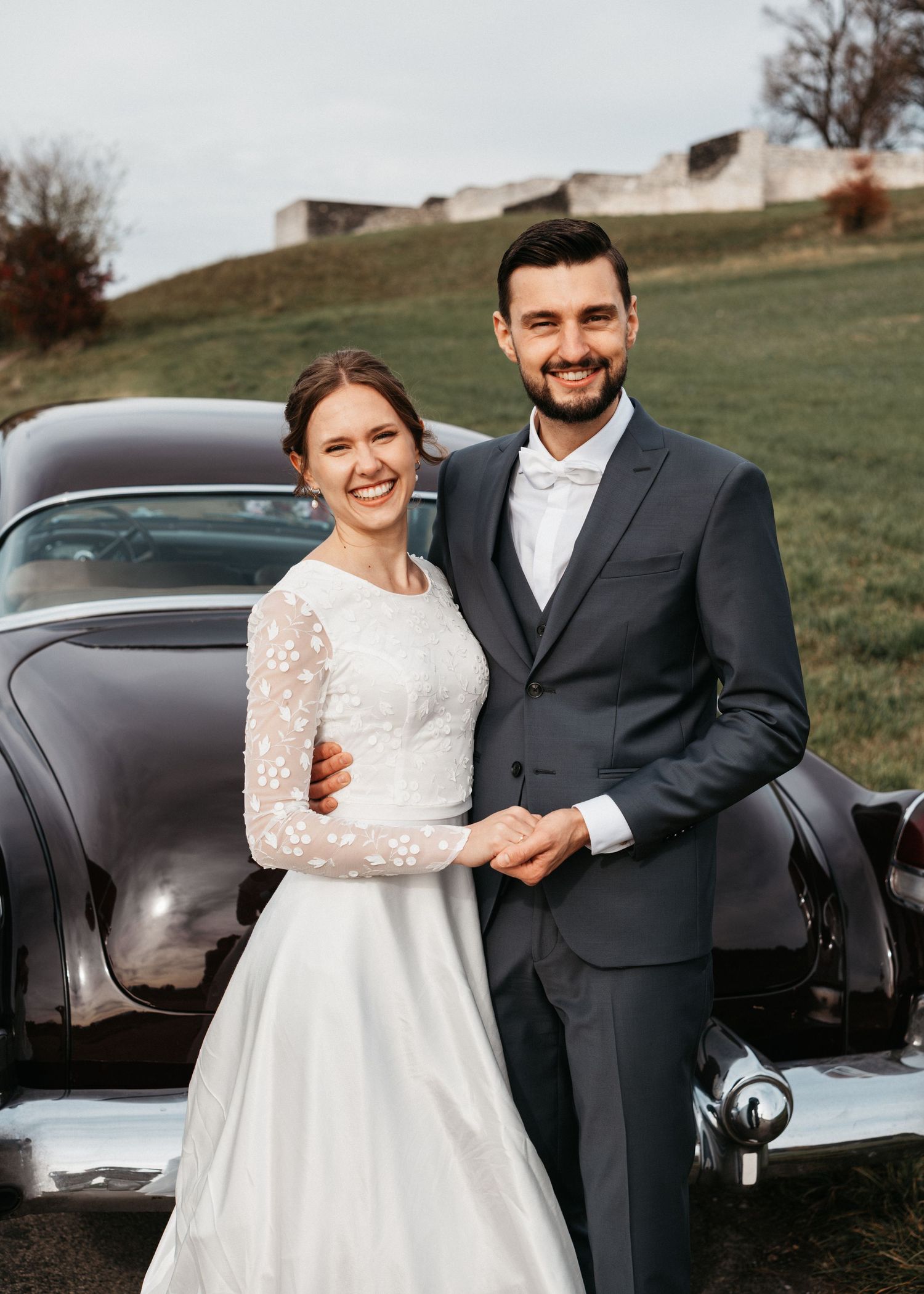 A couple in wedding attire pose together next to a classic black vintage car on a grassy hillside.