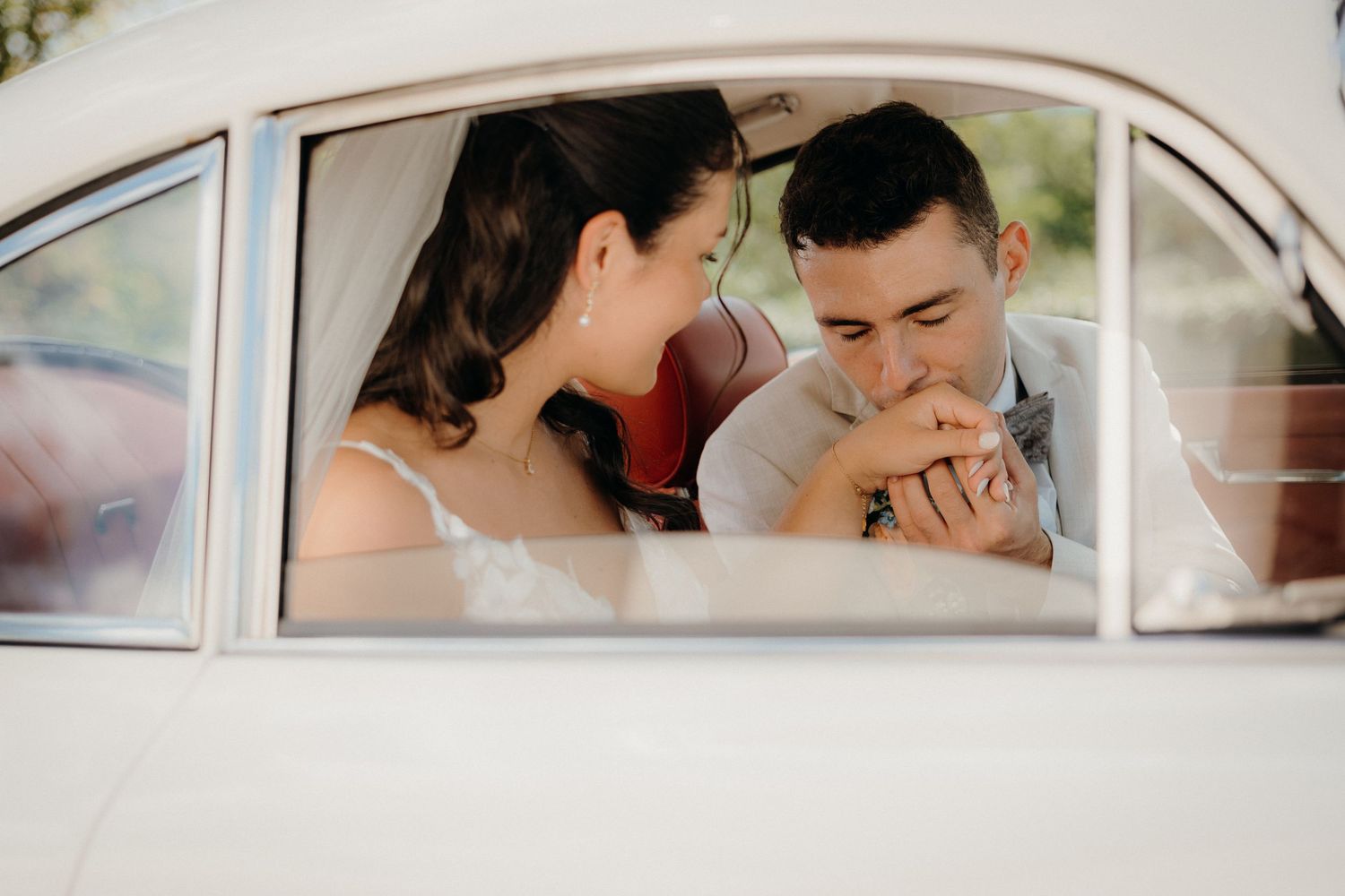 Groom kissing bride's hand tenderly inside a vintage white car on their wedding day.