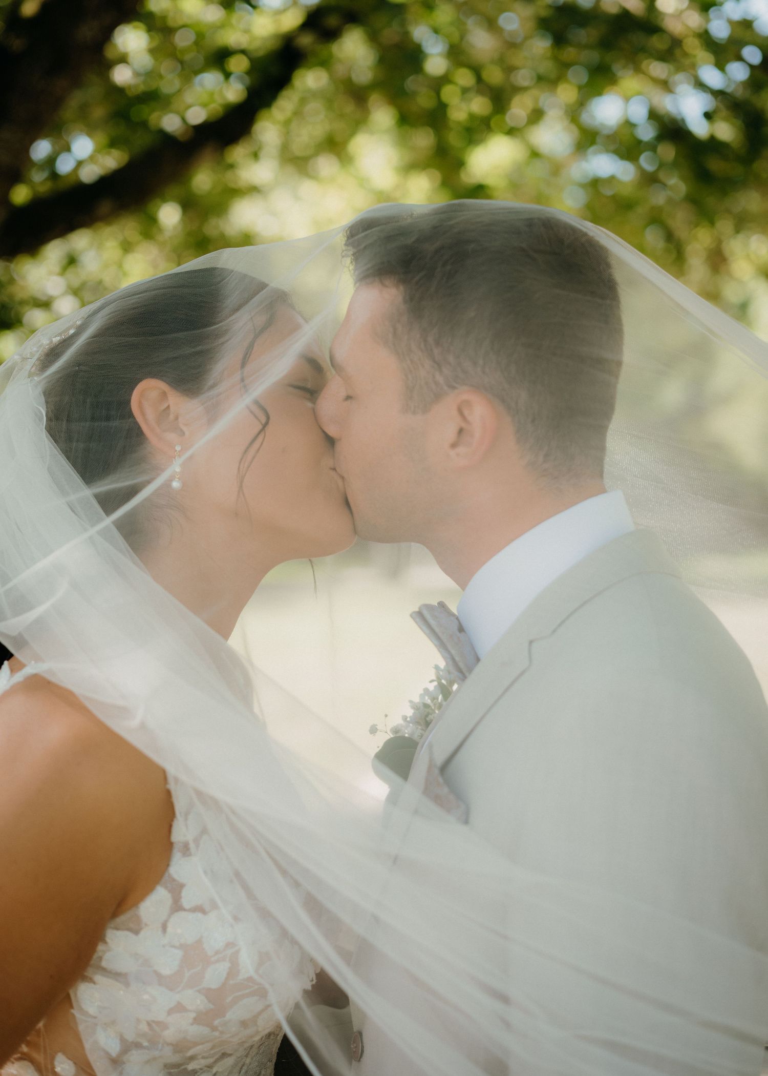 Bride and groom sharing a romantic kiss under a flowing wedding veil outdoors, surrounded by lush green trees.