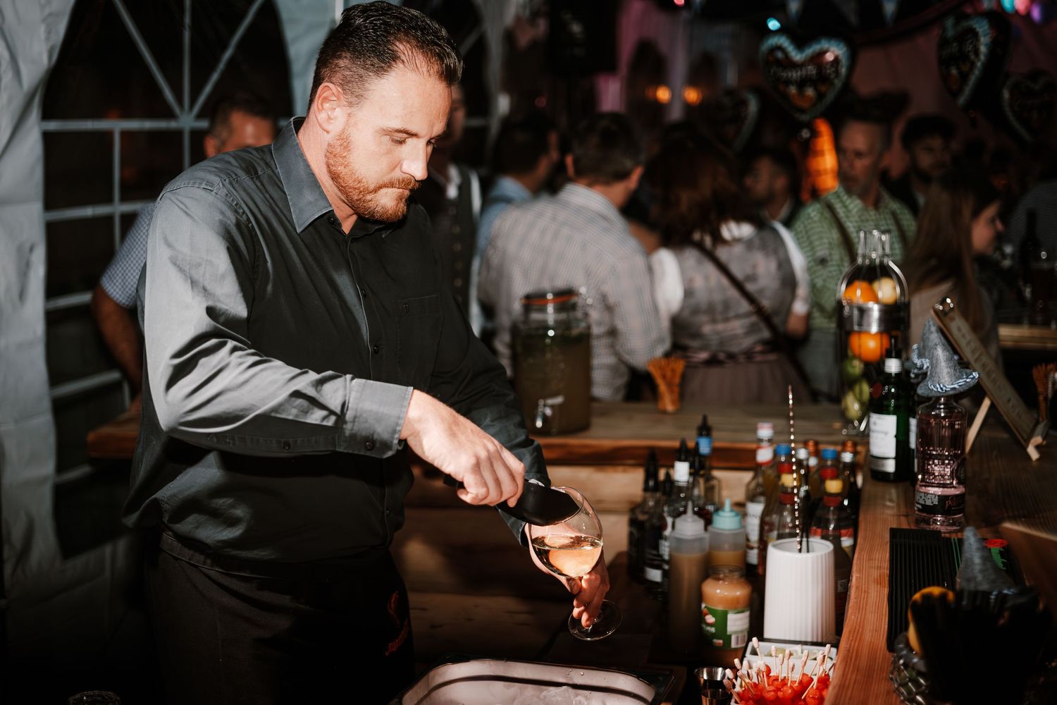 A bartender in a dark dress shirt pours a cocktail at a dimly lit bar with bottles in the background.