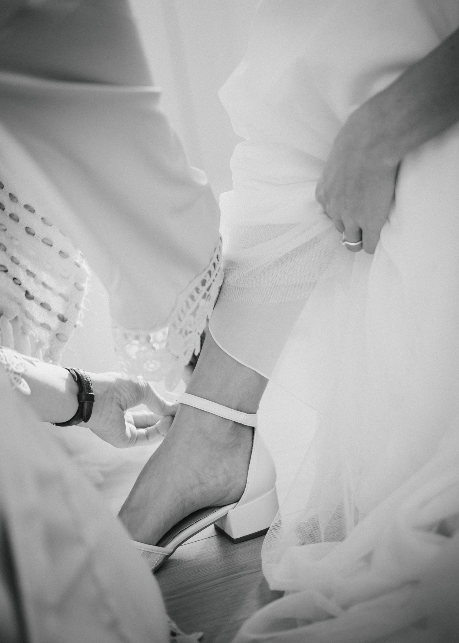 Black and white photo of a bride getting her white wedding shoe strapped onto her ankle by an assistant.