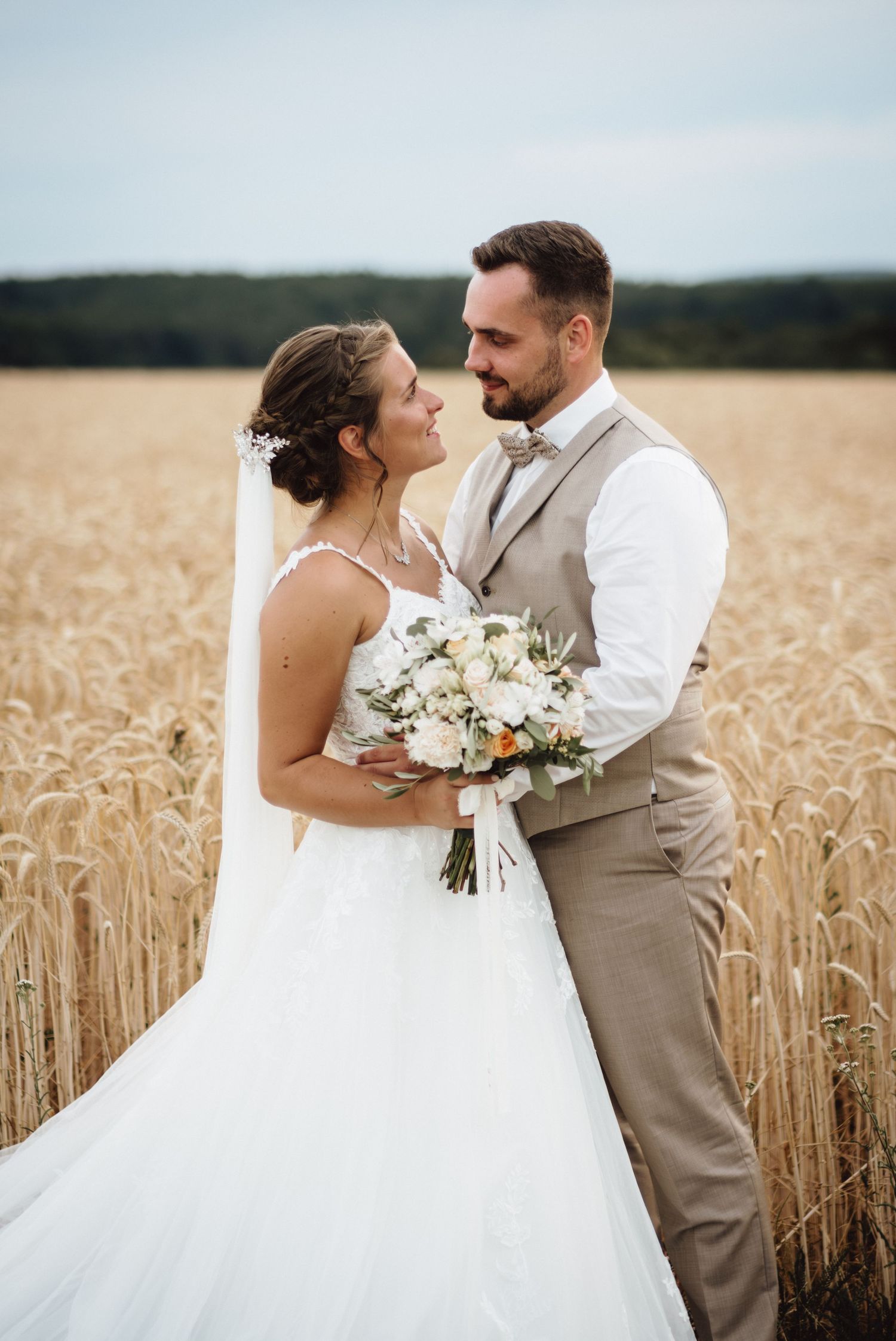 Newlyweds share intimate moments in wheat field, bride in white gown holding floral bouquet.