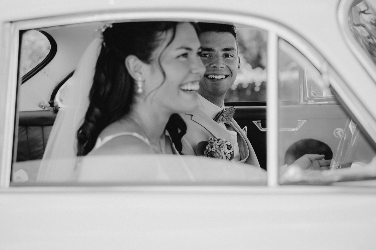 Black and white photo of a smiling bride and groom sitting together in a vintage car on their wedding day.
