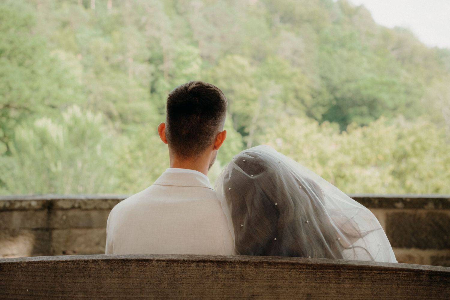 Bride and groom sitting together on a bench from behind, with lush green trees in the background on their wedding day.