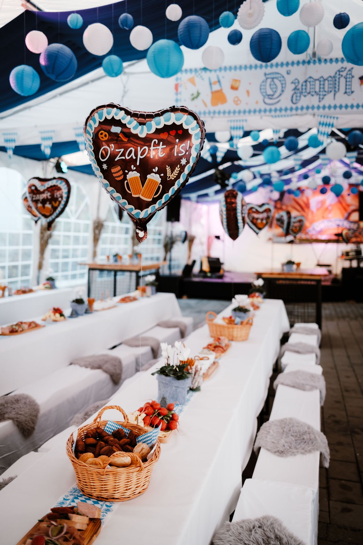 Oktoberfest party setup with heart-shaped balloons, blue decorations, and long white tables with wicker baskets of food.