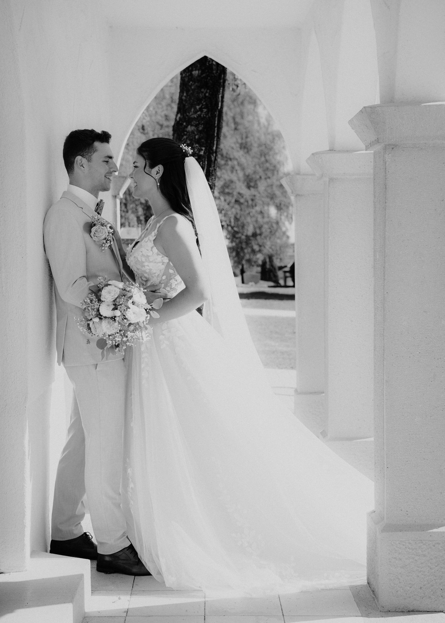 Black and white photo of a bride and groom sharing a romantic moment under an arch, bride holding a bouquet.