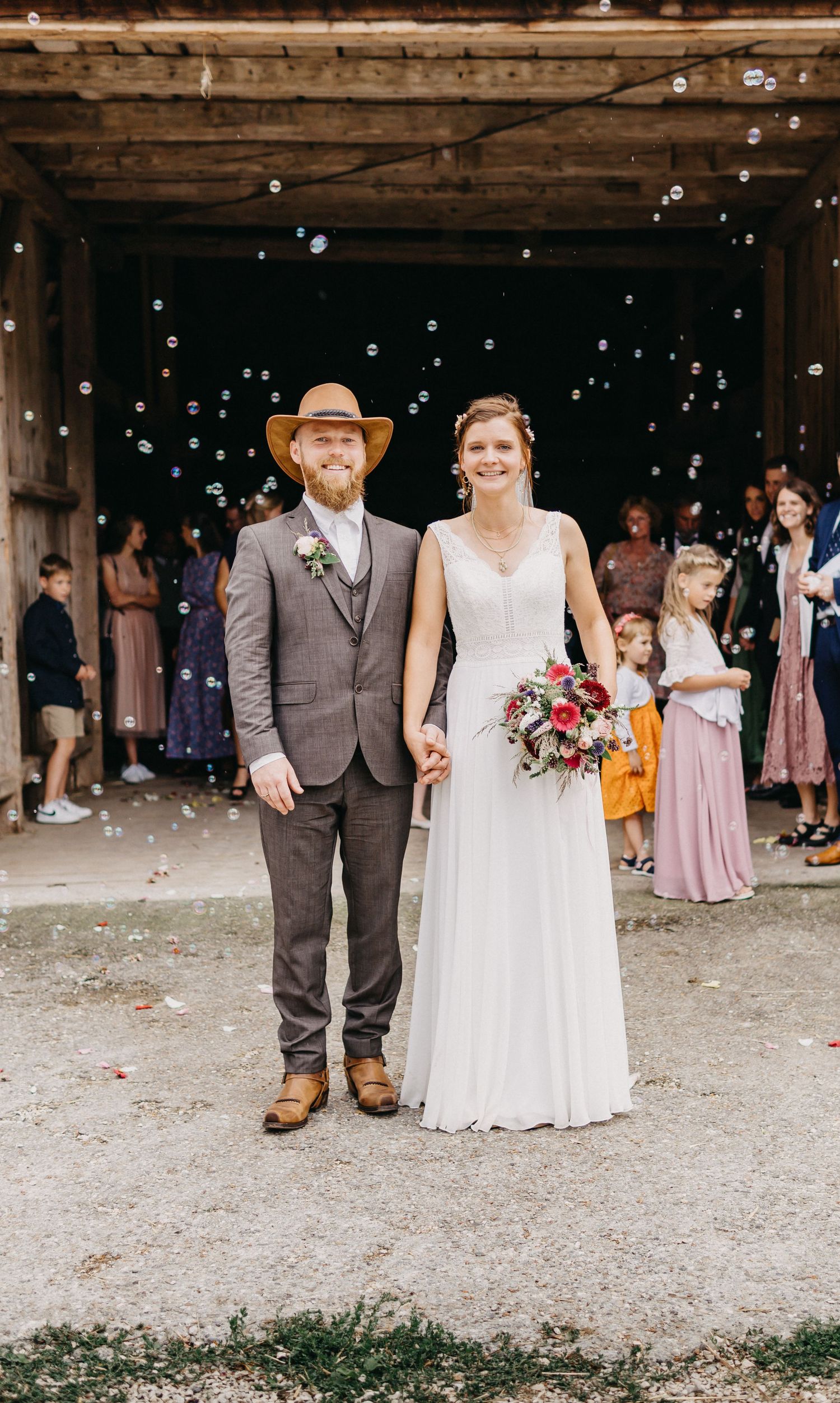 Wedding couple stands together under rustic wooden beams while guests throw confetti during outdoor celebration.