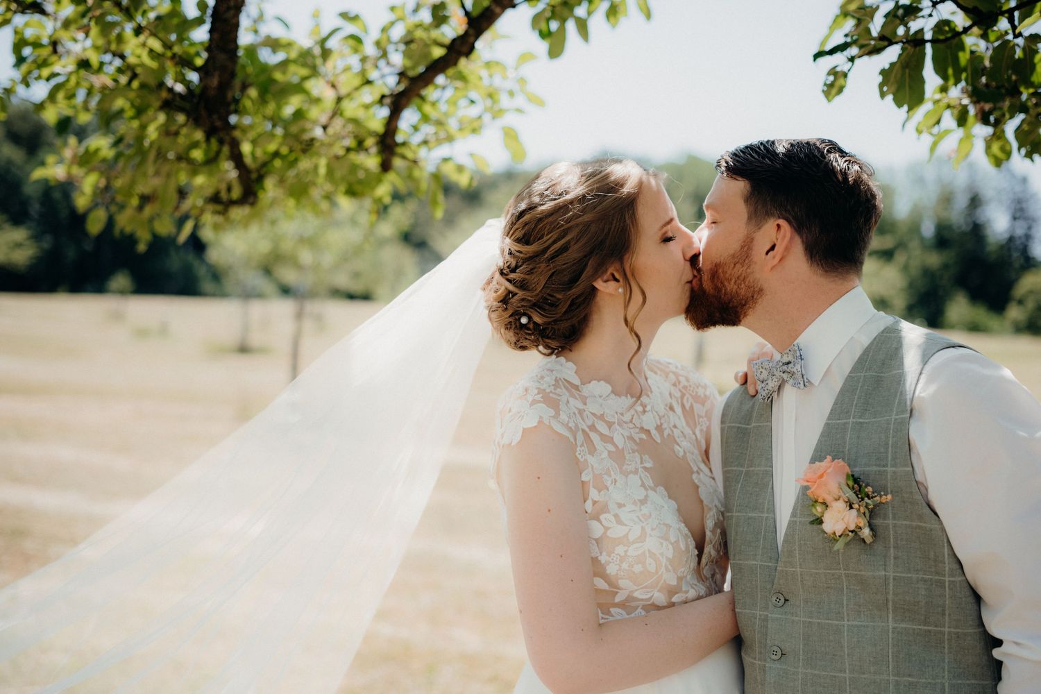 A bride and groom share a romantic kiss outdoors on their wedding day, with a flowing veil and lush green trees.