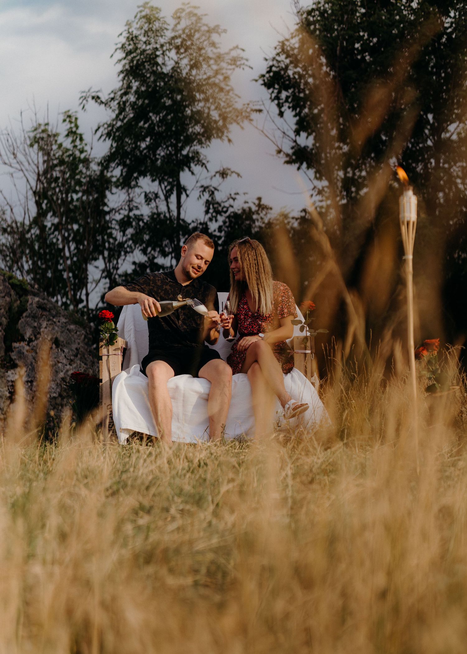 Two people sitting together on a white couch outdoors in a field of tall golden grass at sunset.
