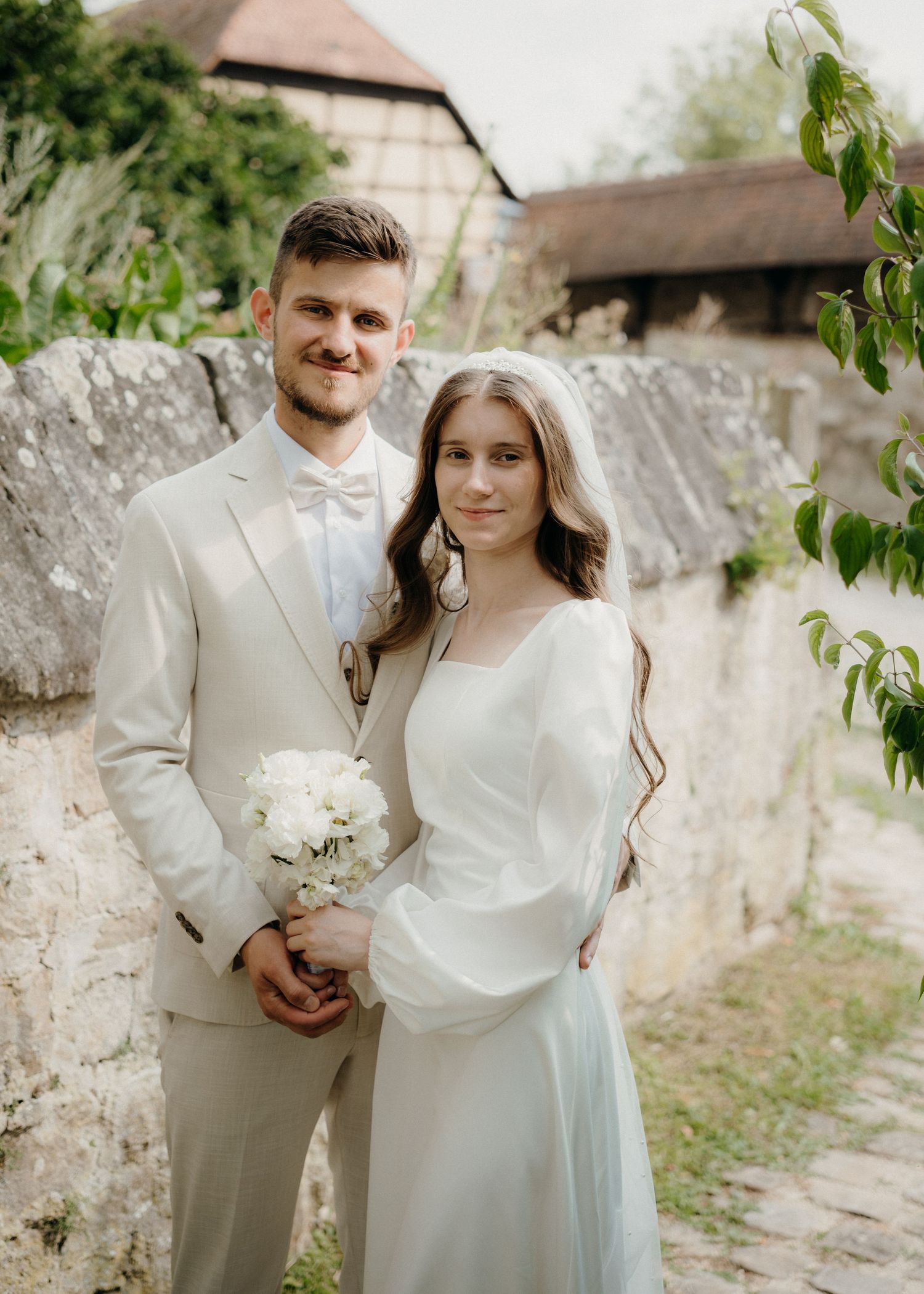 A smiling couple in wedding attire poses outdoors, groom in beige suit, bride in white gown holding a bouquet.