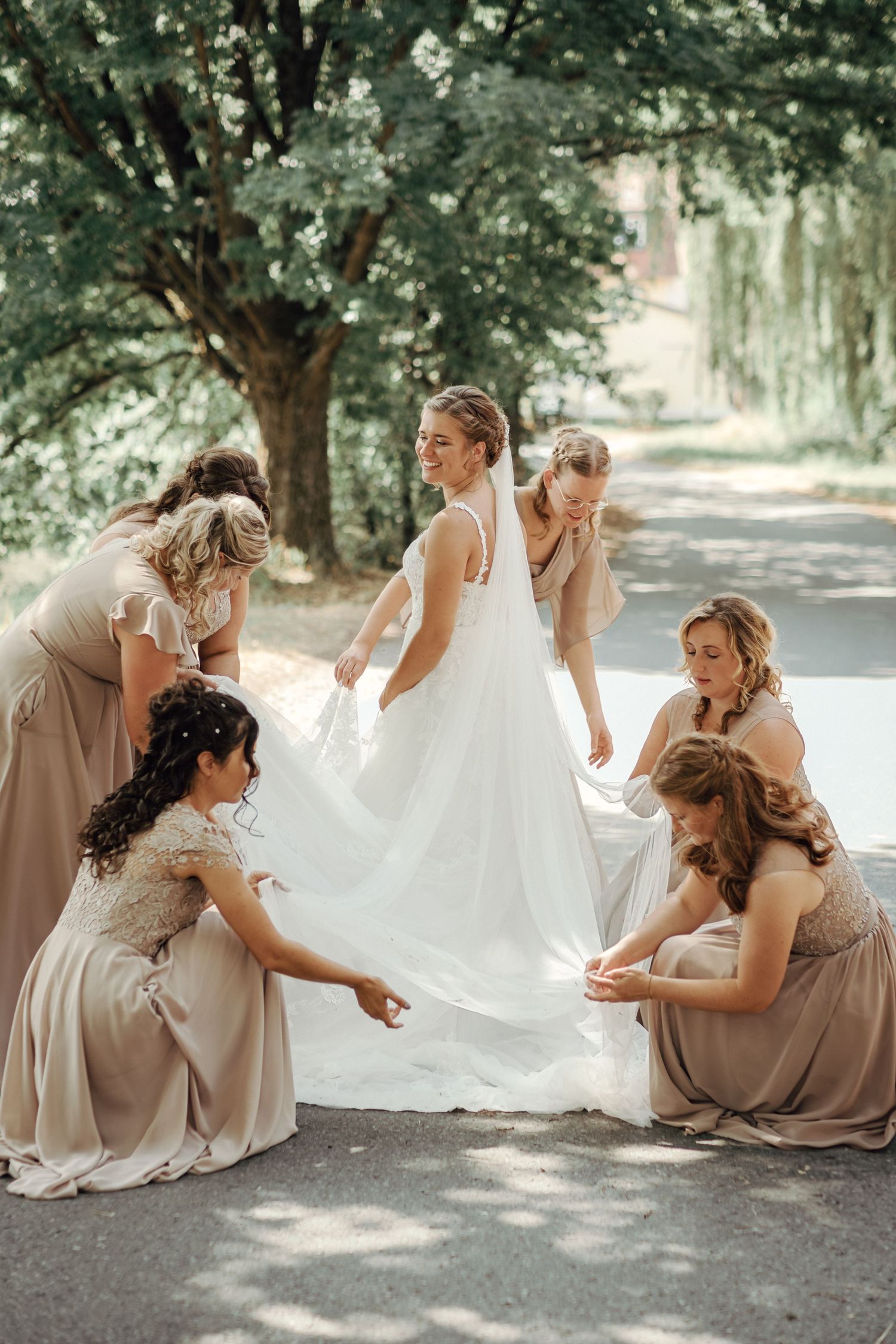 A group of bridesmaids in beige dresses help arrange the bride's white wedding gown train on a garden path.