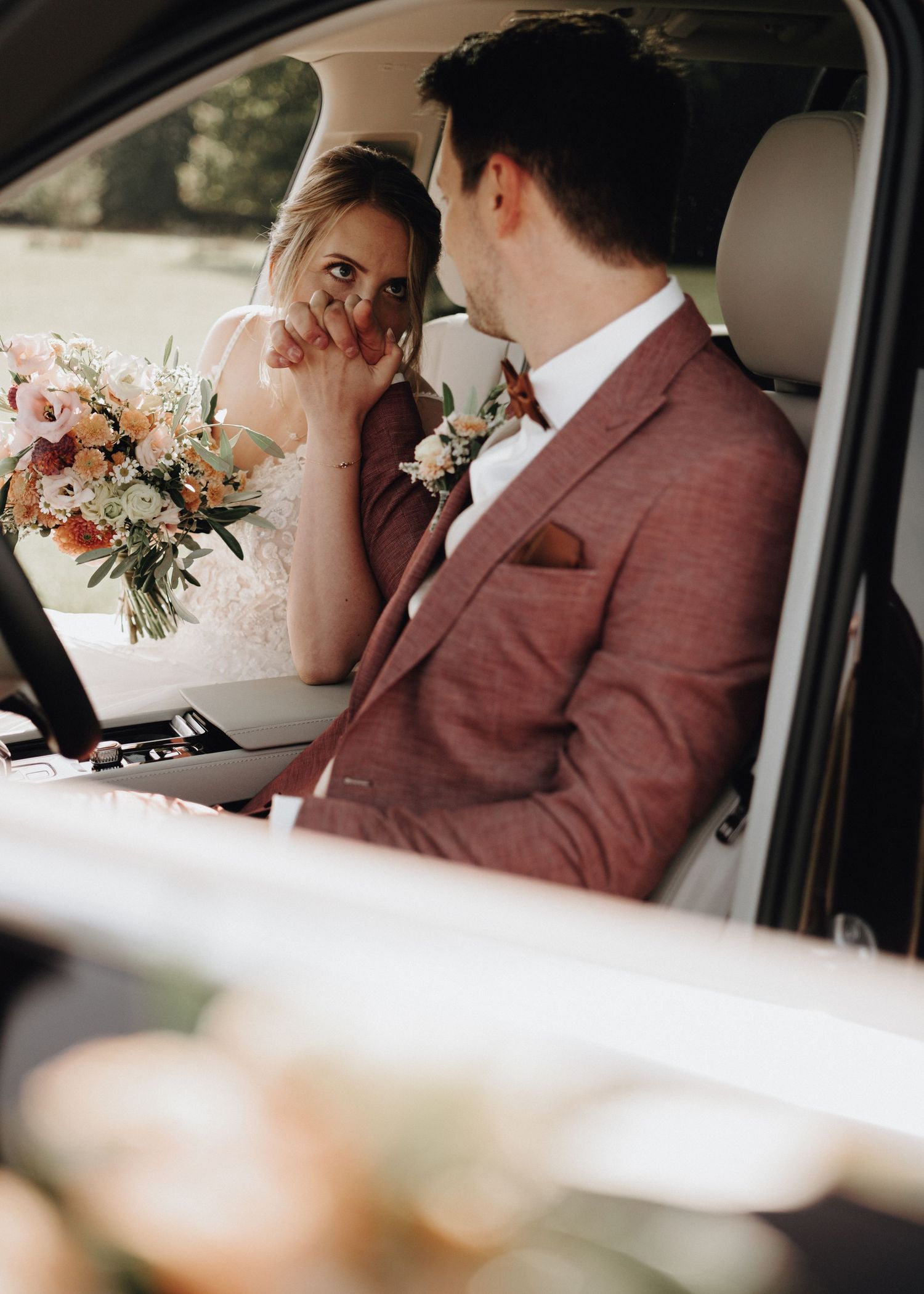 Newlywed couple shares intimate moments together in vintage car backseat with elegant floral bouquet.