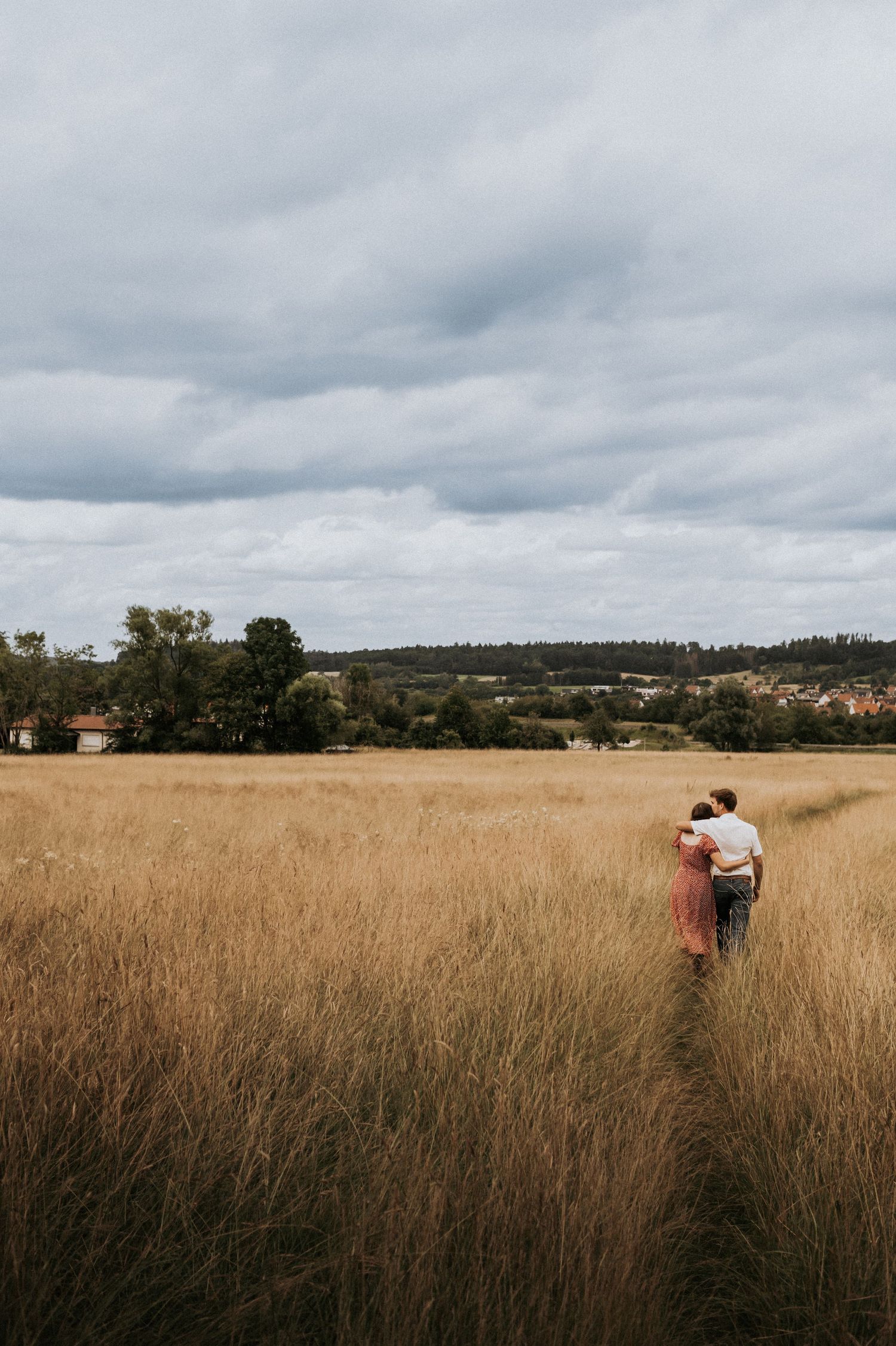 A couple walks through a golden wheat field under cloudy skies in a romantic countryside setting.