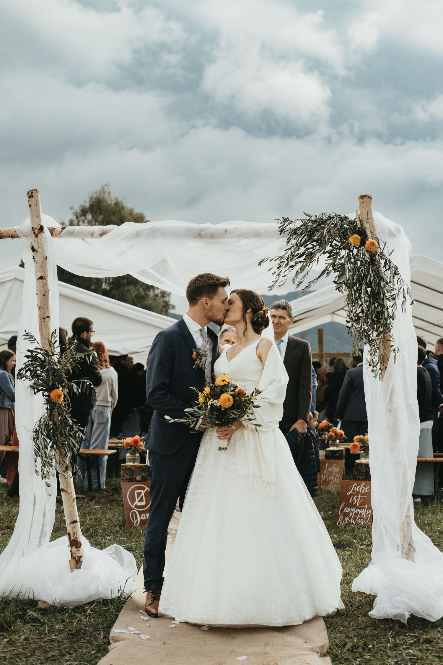Outdoor wedding ceremony with white drapes and greenery decorations under cloudy skies.