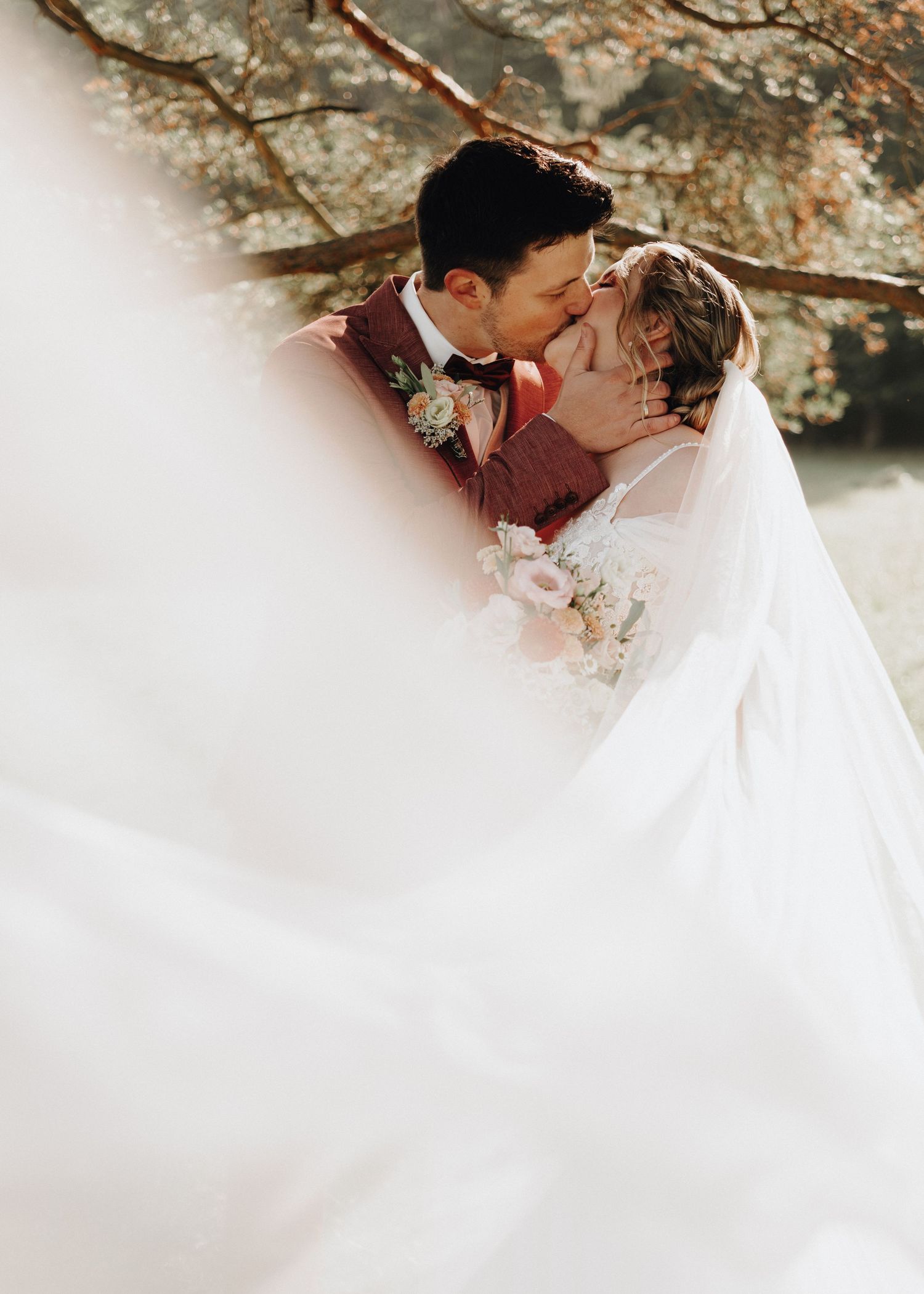 A romantic couple shares a tender kiss under autumn trees with a dreamy white veil flowing in the wind.