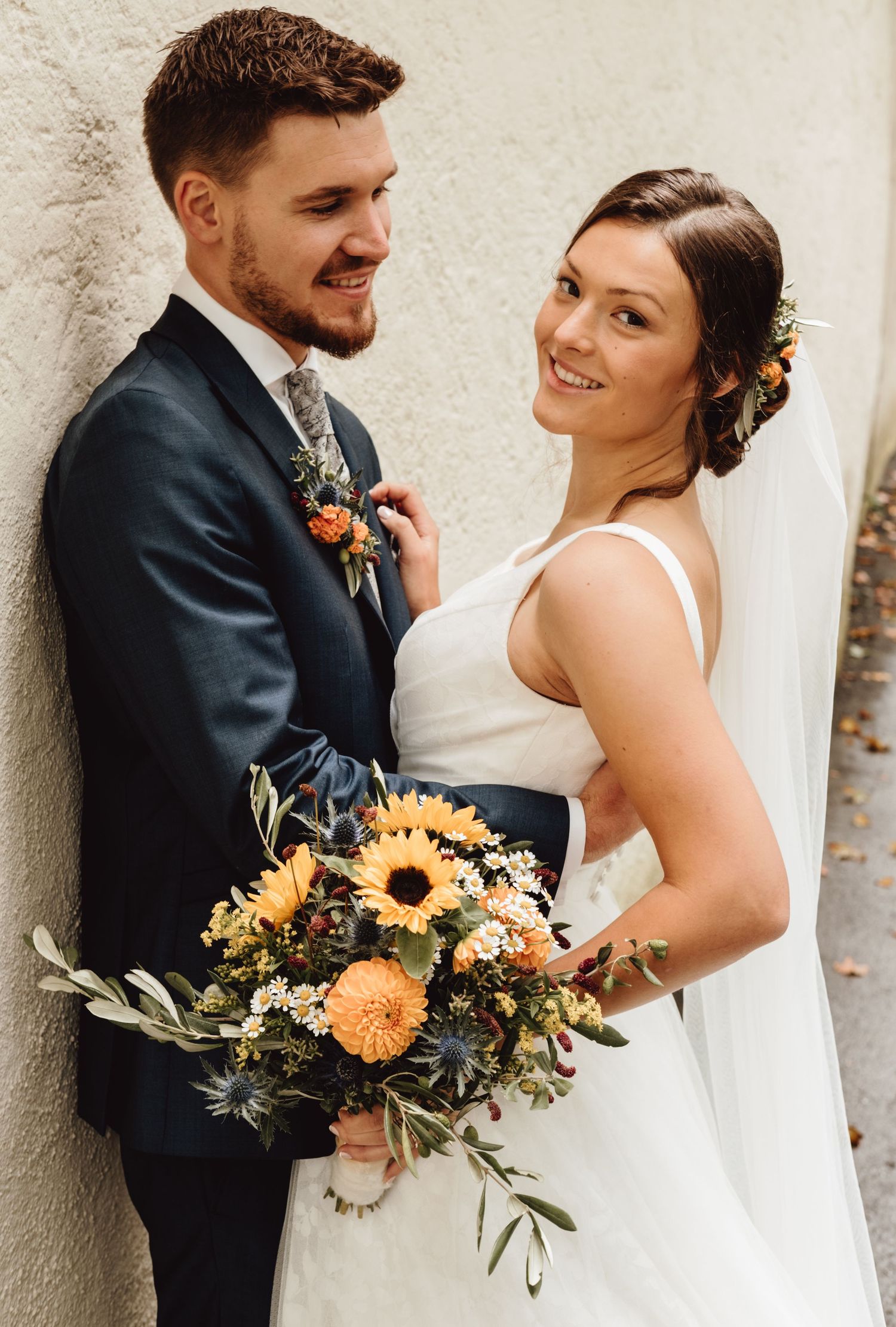 A romantic wedding couple poses against a light stone wall with sunflowers and orange blooms in their bouquet.