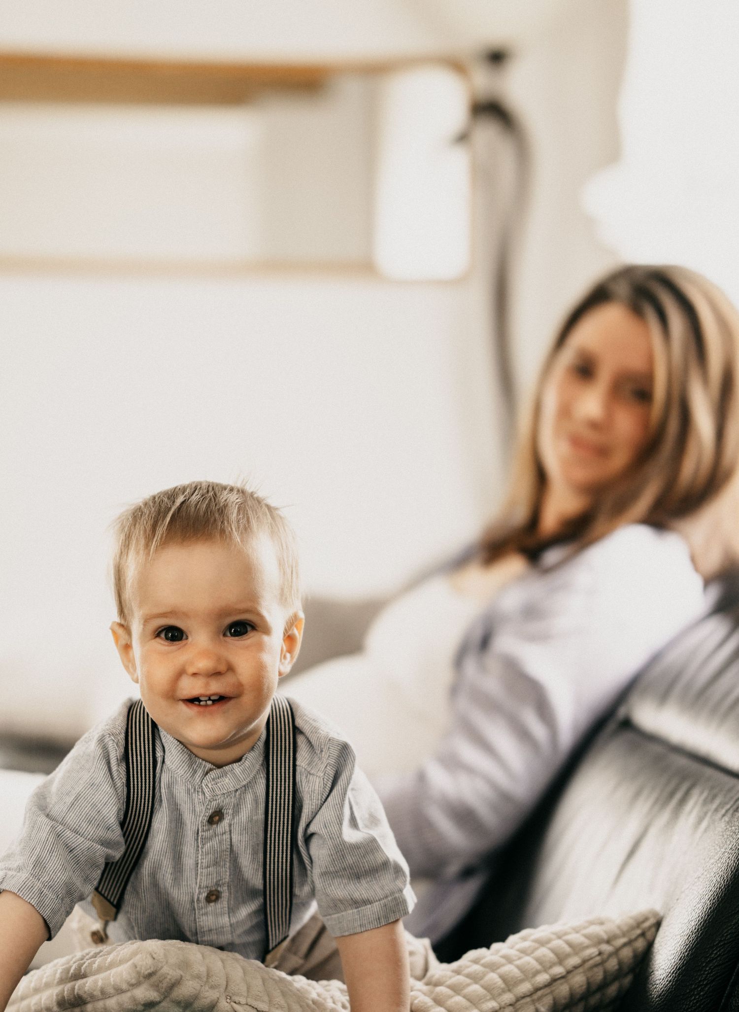 Baby in gray shirt and suspenders smiles while sitting on bed in bright room.