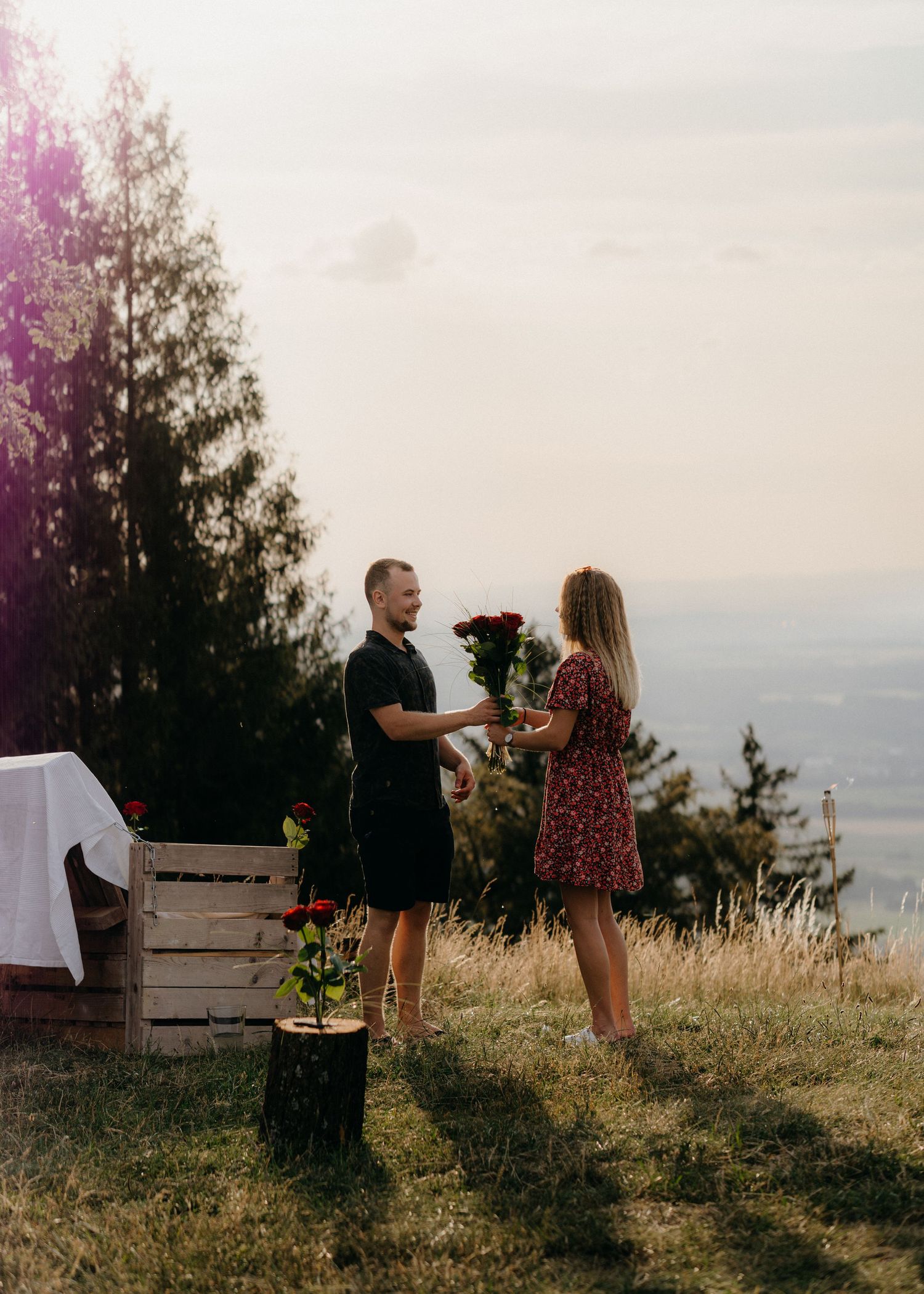A couple shares a tender moment at sunset on a grassy hilltop overlooking a scenic landscape.
