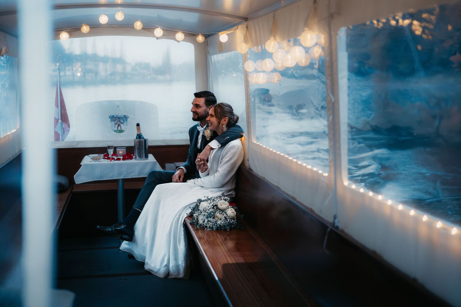 A scenic wedding venue with string lights and water views visible through large windows at sunset.