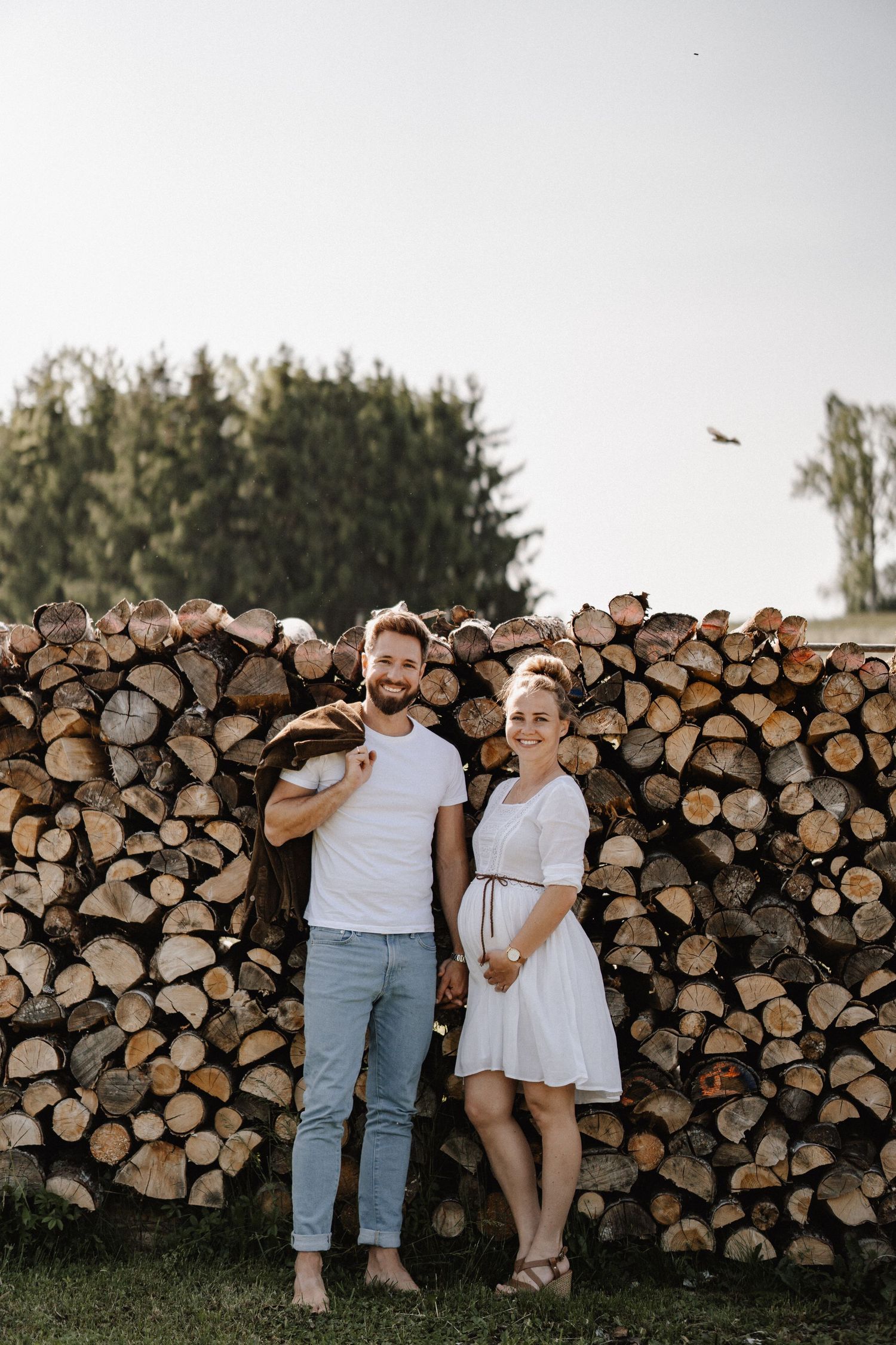 Couple stands together in front of stacked firewood logs during an outdoor photo session at sunset.