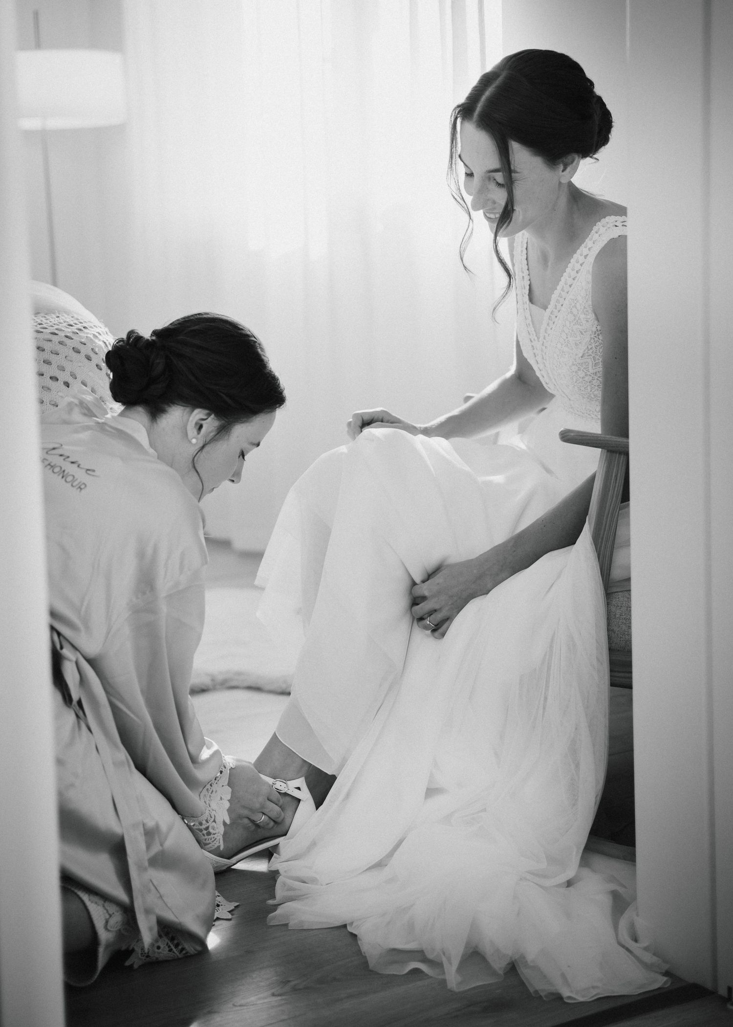 Black and white photo of a bride in white gown being helped with her shoes by a bridesmaid before the wedding.