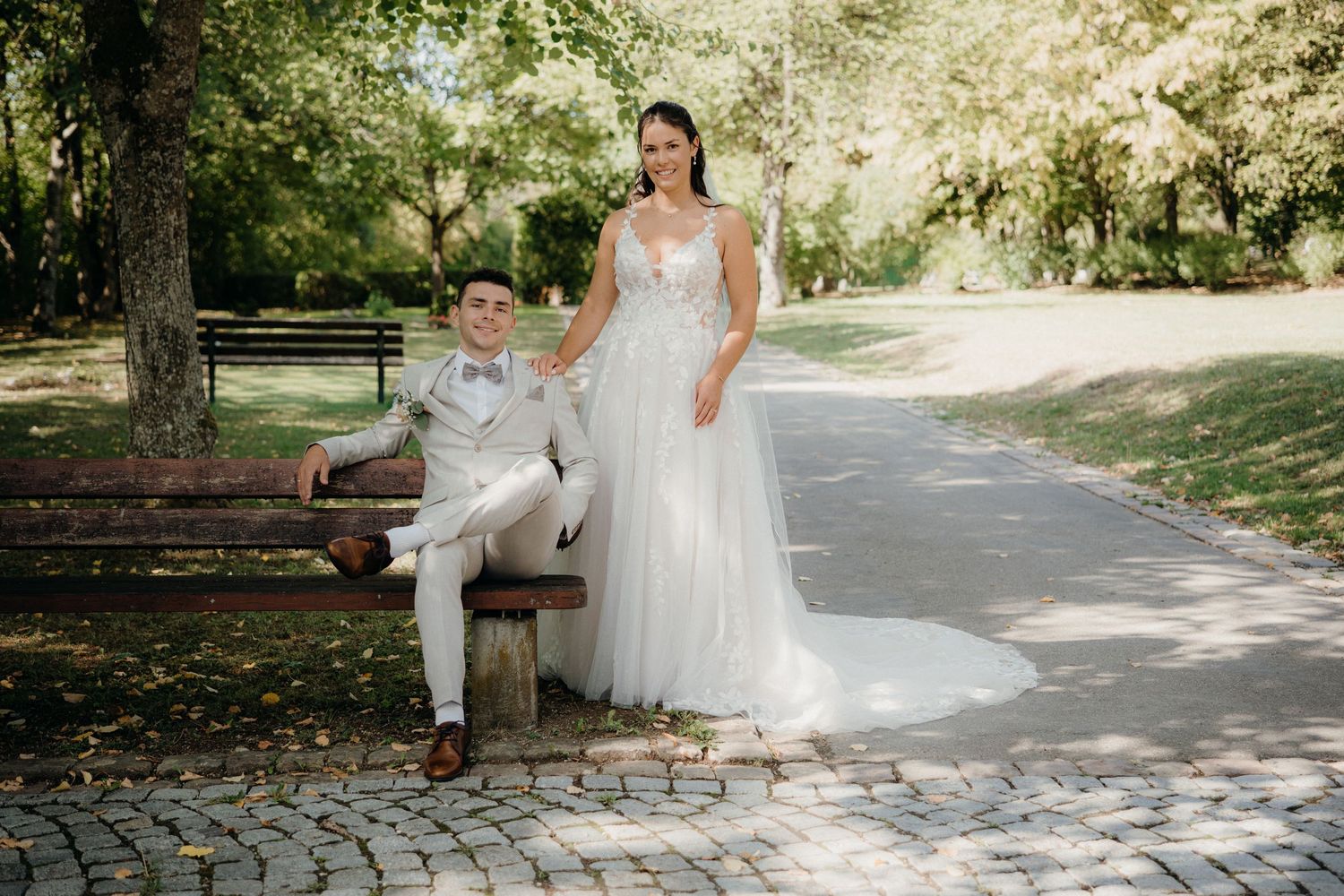Bride in white gown stands beside groom in gray suit seated on park bench, surrounded by lush green trees.