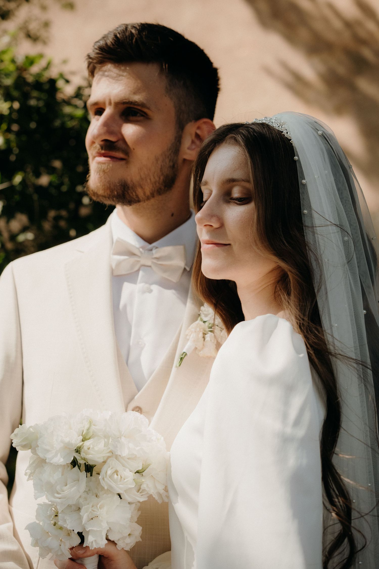 Bride and groom in white wedding attire pose together, bride holding white bouquet and wearing veil.