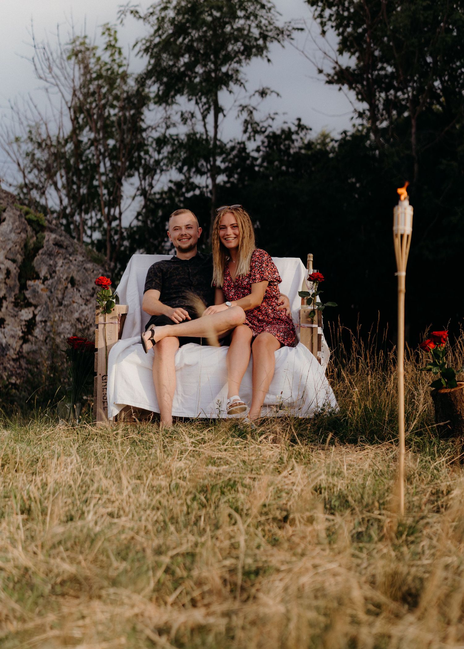 A couple lounges on a white couch outdoors in a grassy field at dusk with tiki torches creating a romantic ambiance.