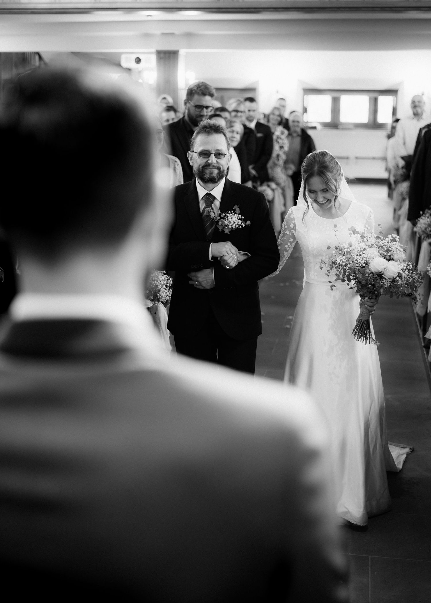 A black and white wedding ceremony photo shows a bride walking down the aisle in a traditional white gown.