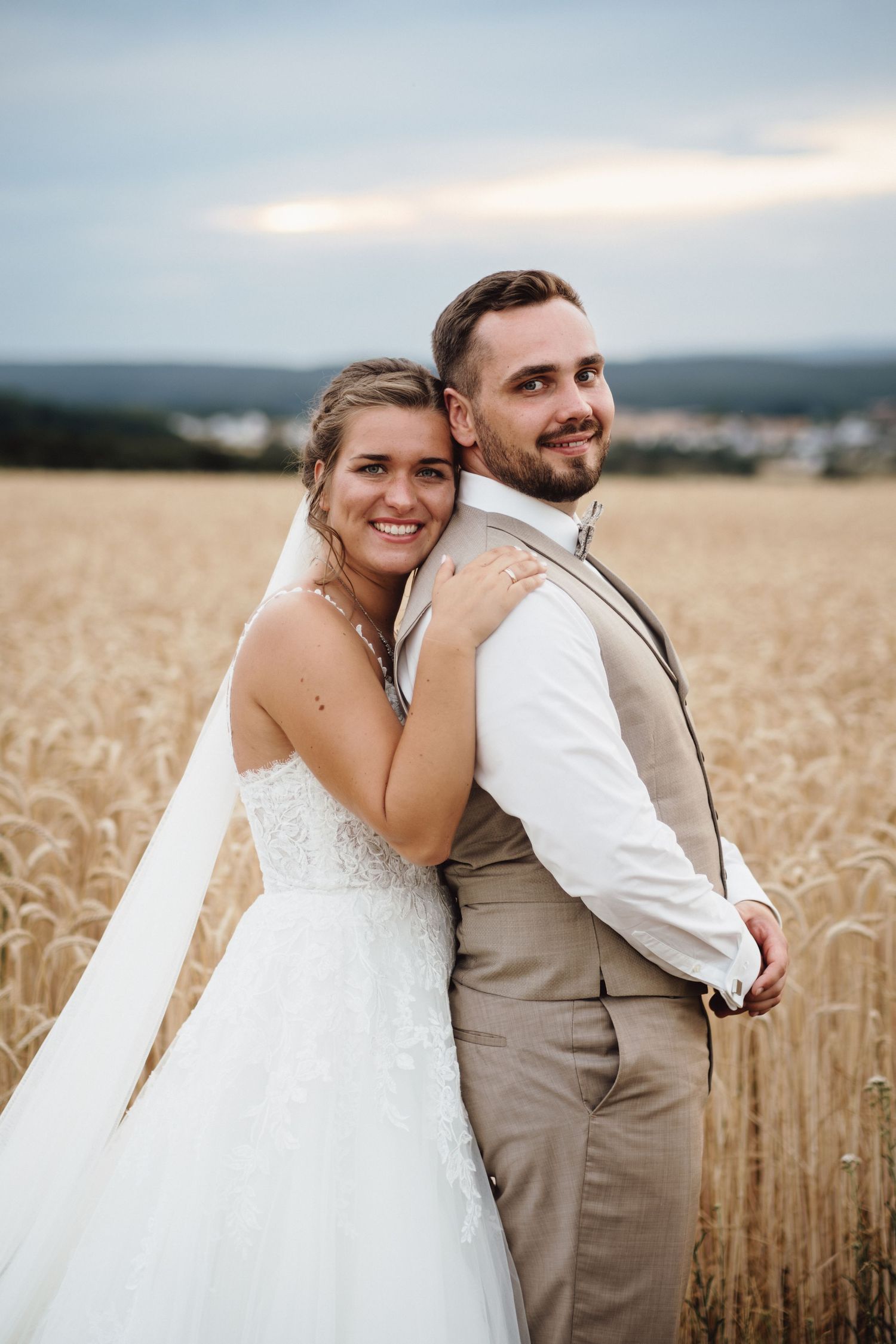 A couple poses together in a golden wheat field at sunset wearing wedding attire.