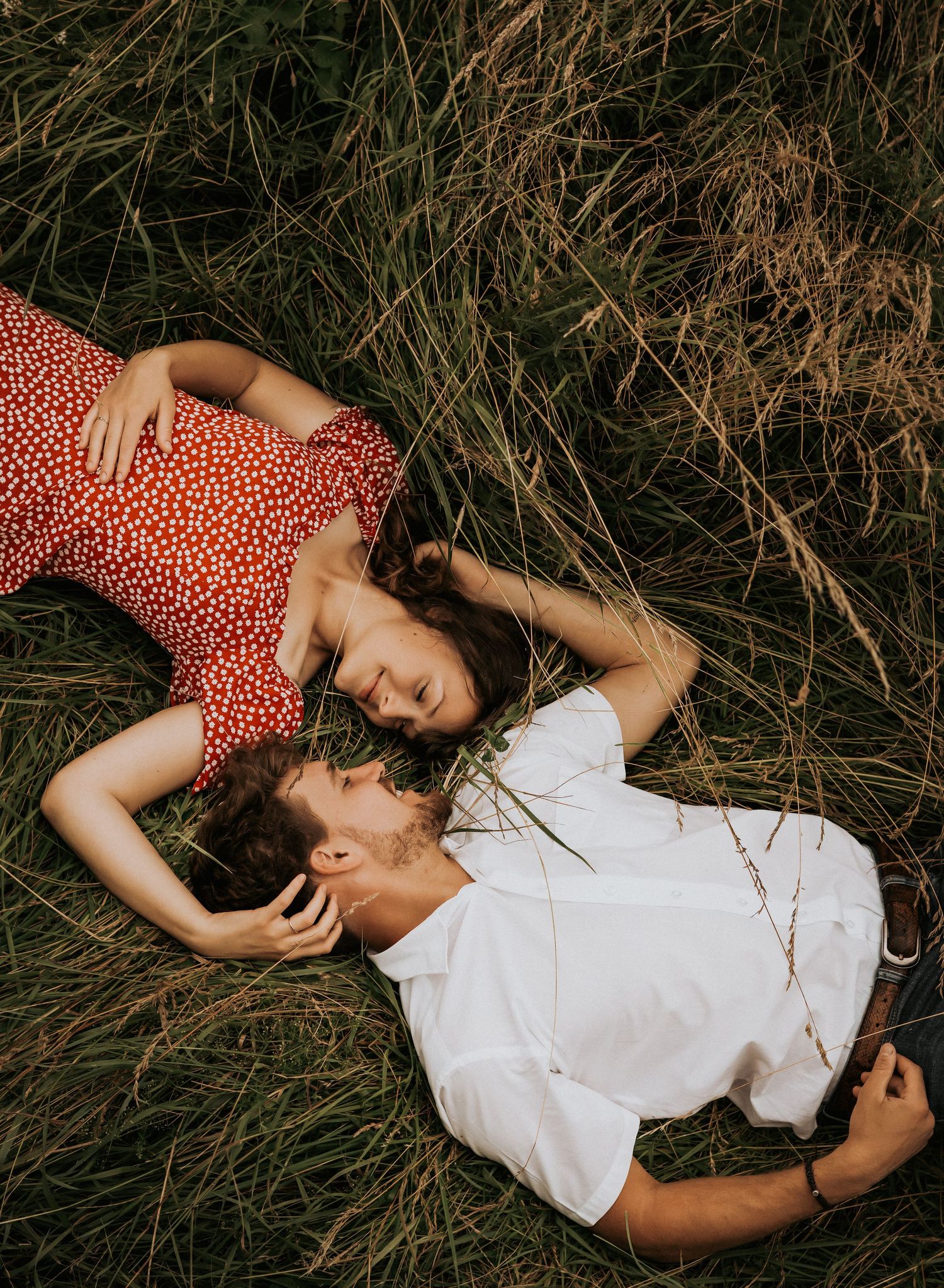 A couple in red and white outfits lying together in a field of tall grass during a romantic photoshoot.
