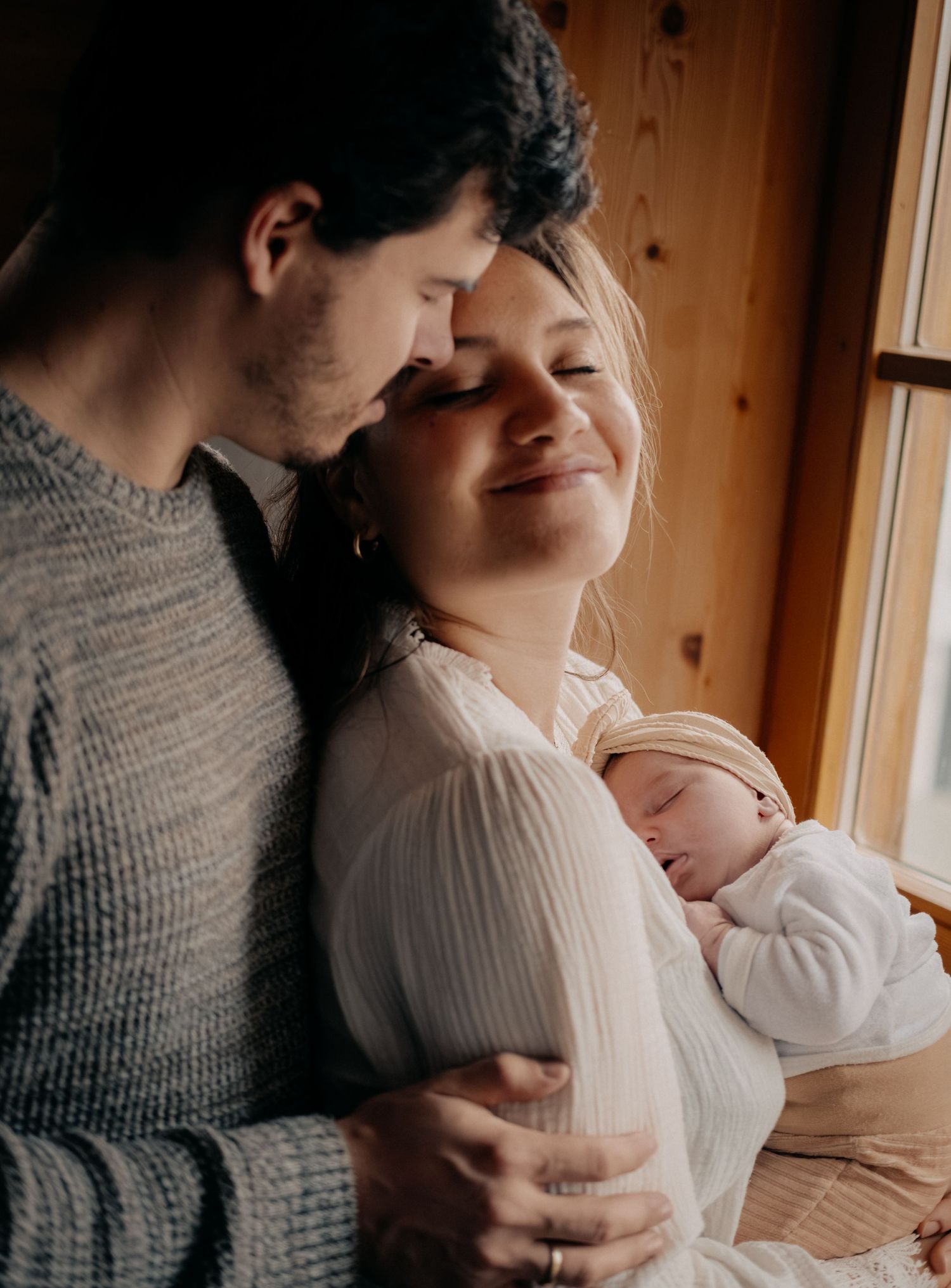 A tender moment between parents and their newborn baby in a sunlit wooden room.