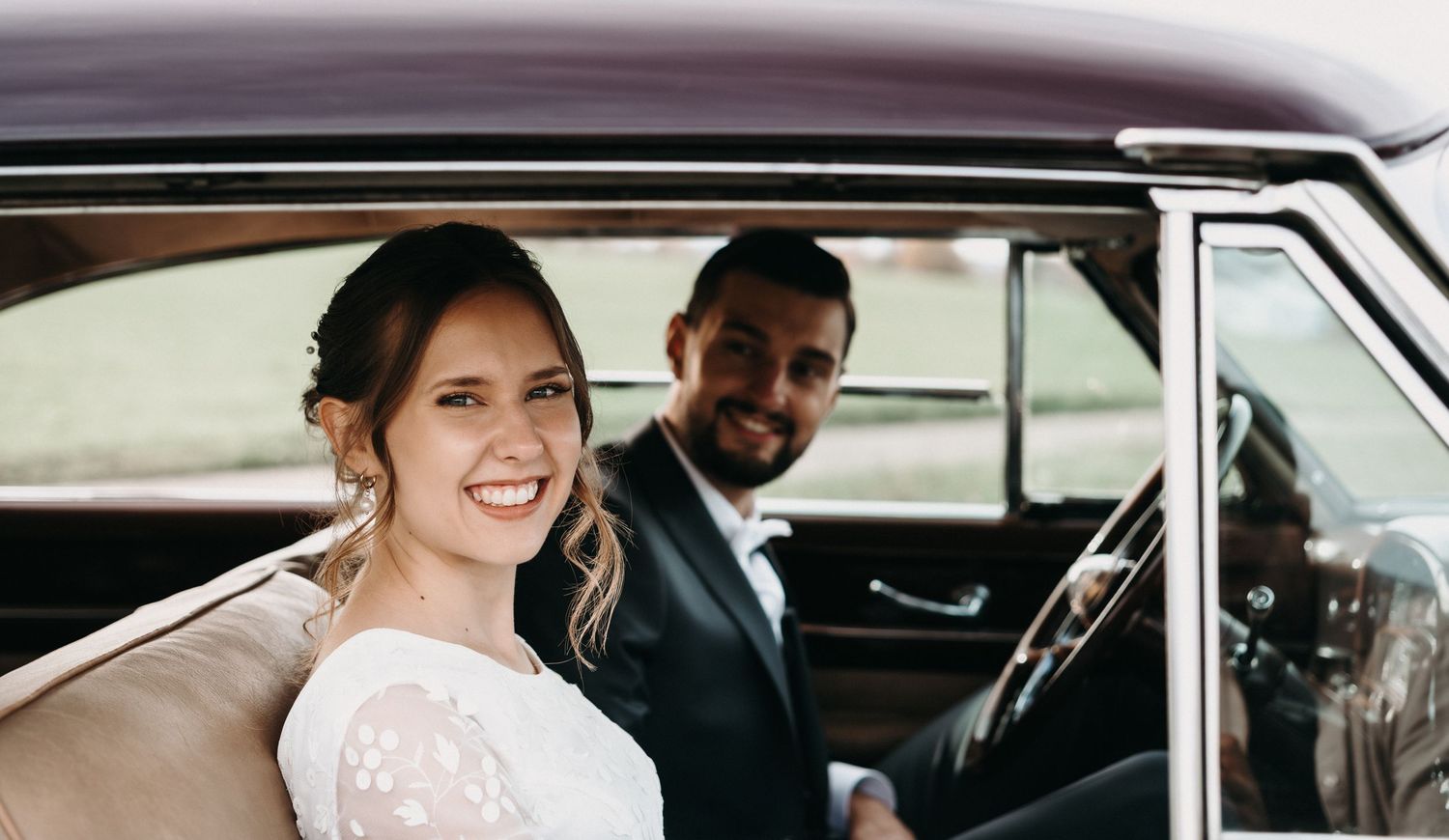 A happy couple sits together in a vintage car on their wedding day, with the bride wearing a white lace dress.