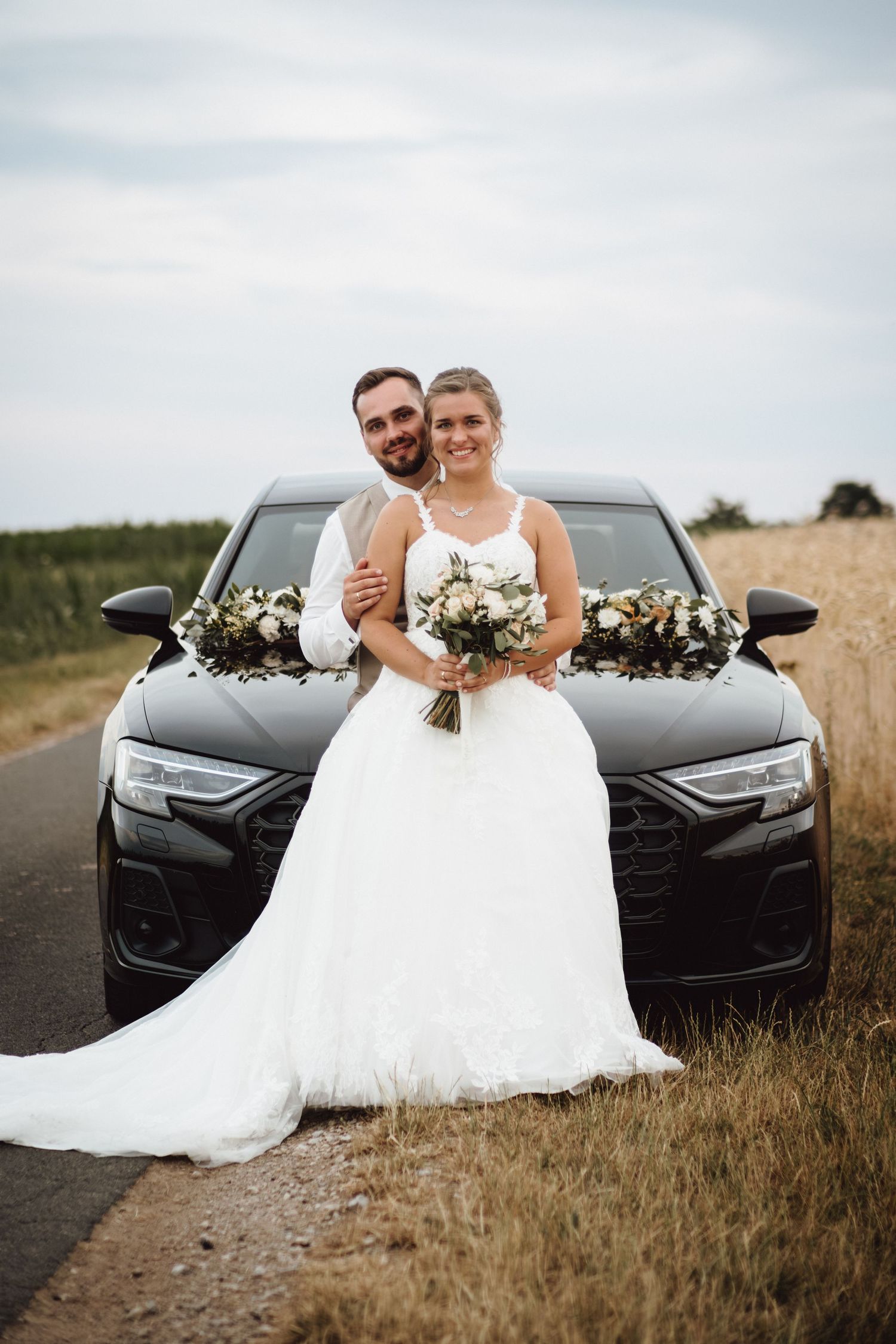 A newlywed couple poses in front of a black car decorated with white floral arrangements in a rural field setting.