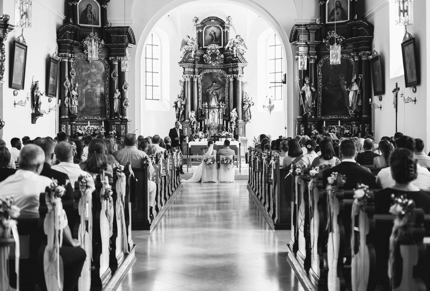 A traditional church wedding ceremony with guests seated in pews along a white aisle leading to an ornate altar.